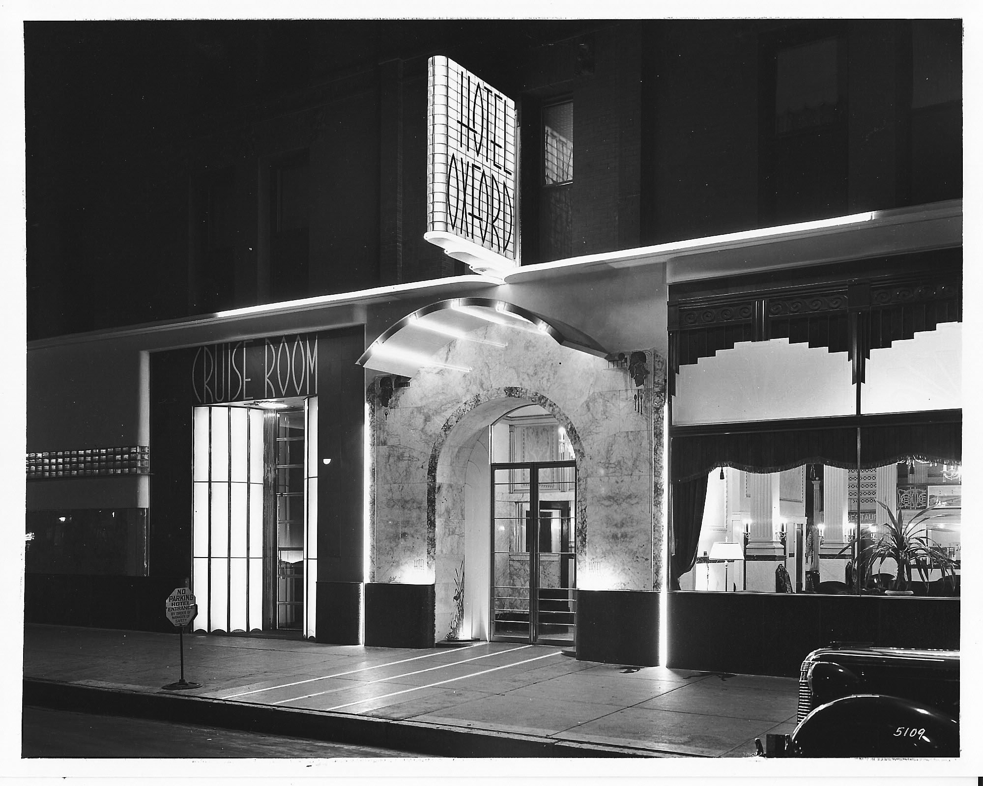 Black and white nighttime photo of Hotel Oxford entrance with lit arched doorway and neon-lit "Cruise Room" sign to the left, a window showing interior lamps and curtains to the right.