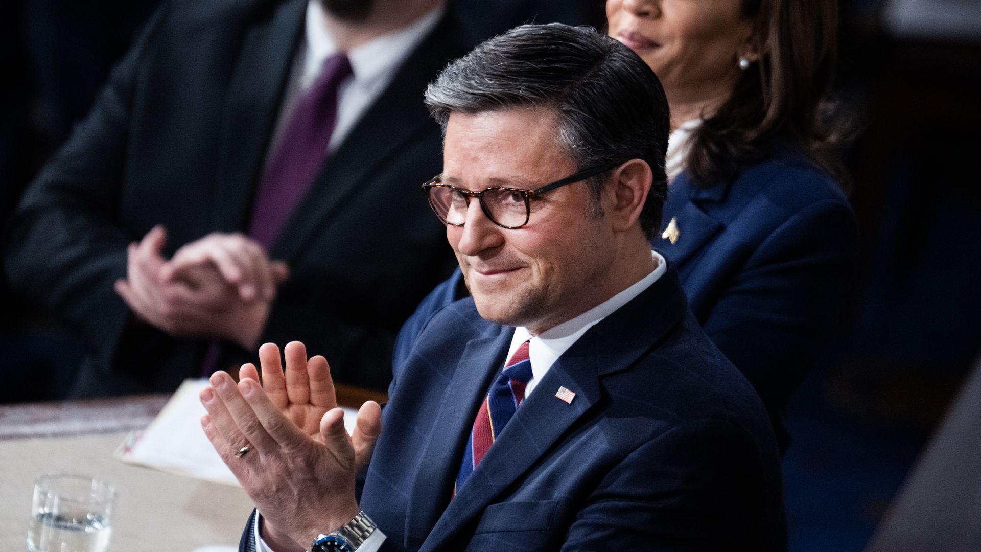 House Speaker Mike Johnson, wearing a blue suit, white shirt, red and blue tie and glasses, clapping in the House chamber.