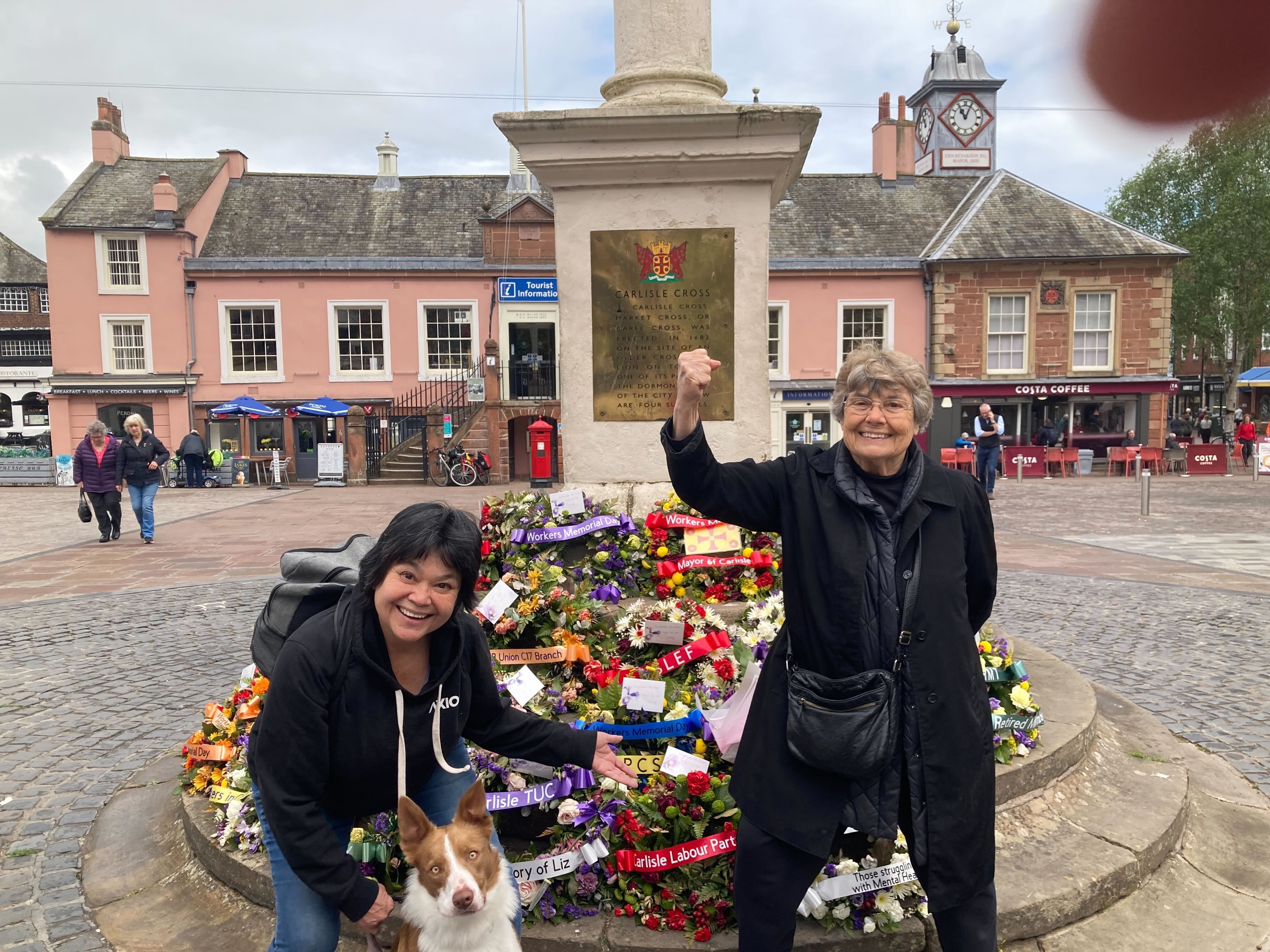 Two smiling women pose by a memorial pedestal in a town square at Carlisle Cross, with a flower wreath and a dog nearby. Pink historic buildings, a clock tower, and Costa Coffee in the background.