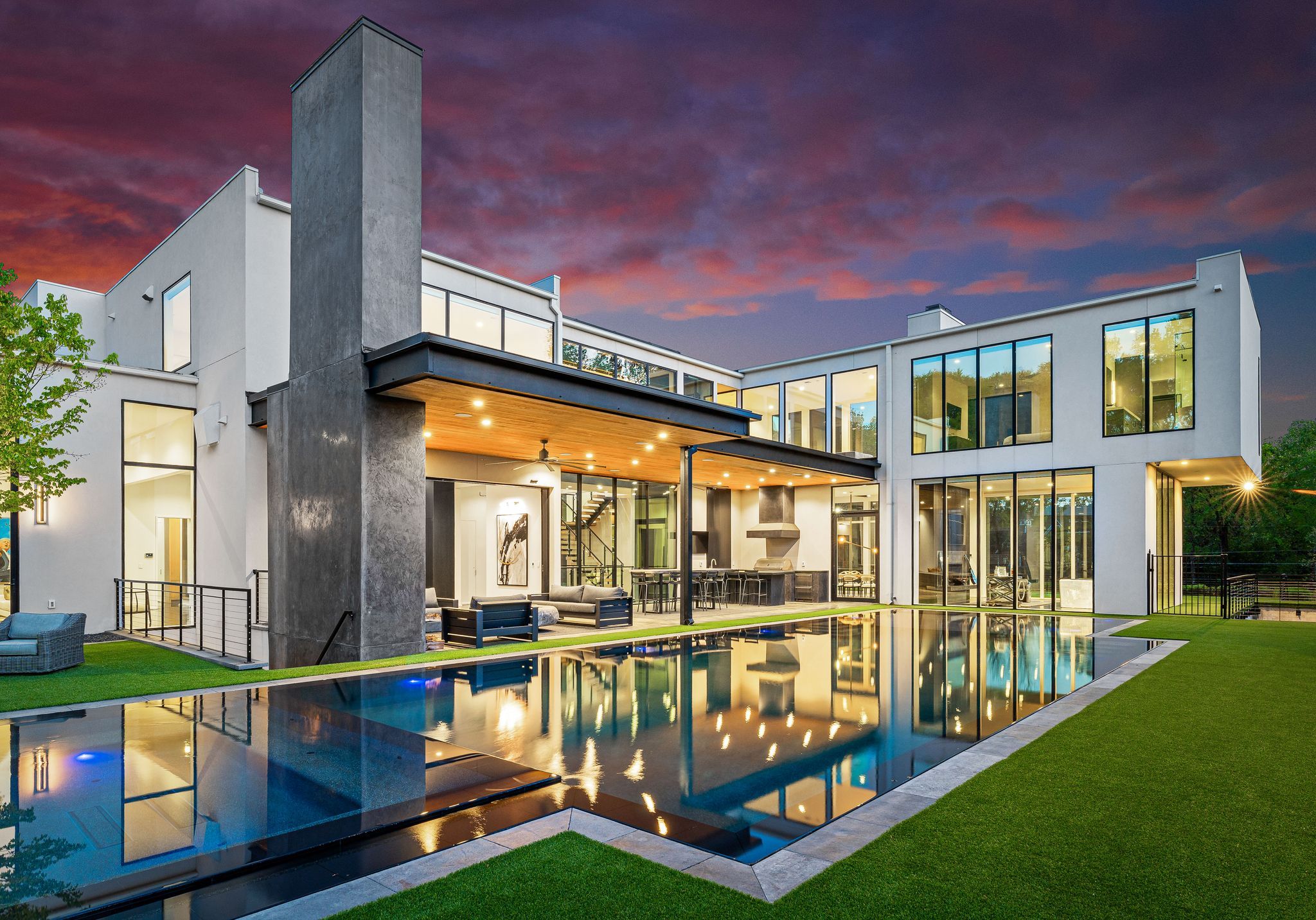 Modern white two-story house with large glass windows, outdoor lounge, and kitchen area overlooking a rectangular illuminated pool, set against a vibrant purple and pink sunset sky.