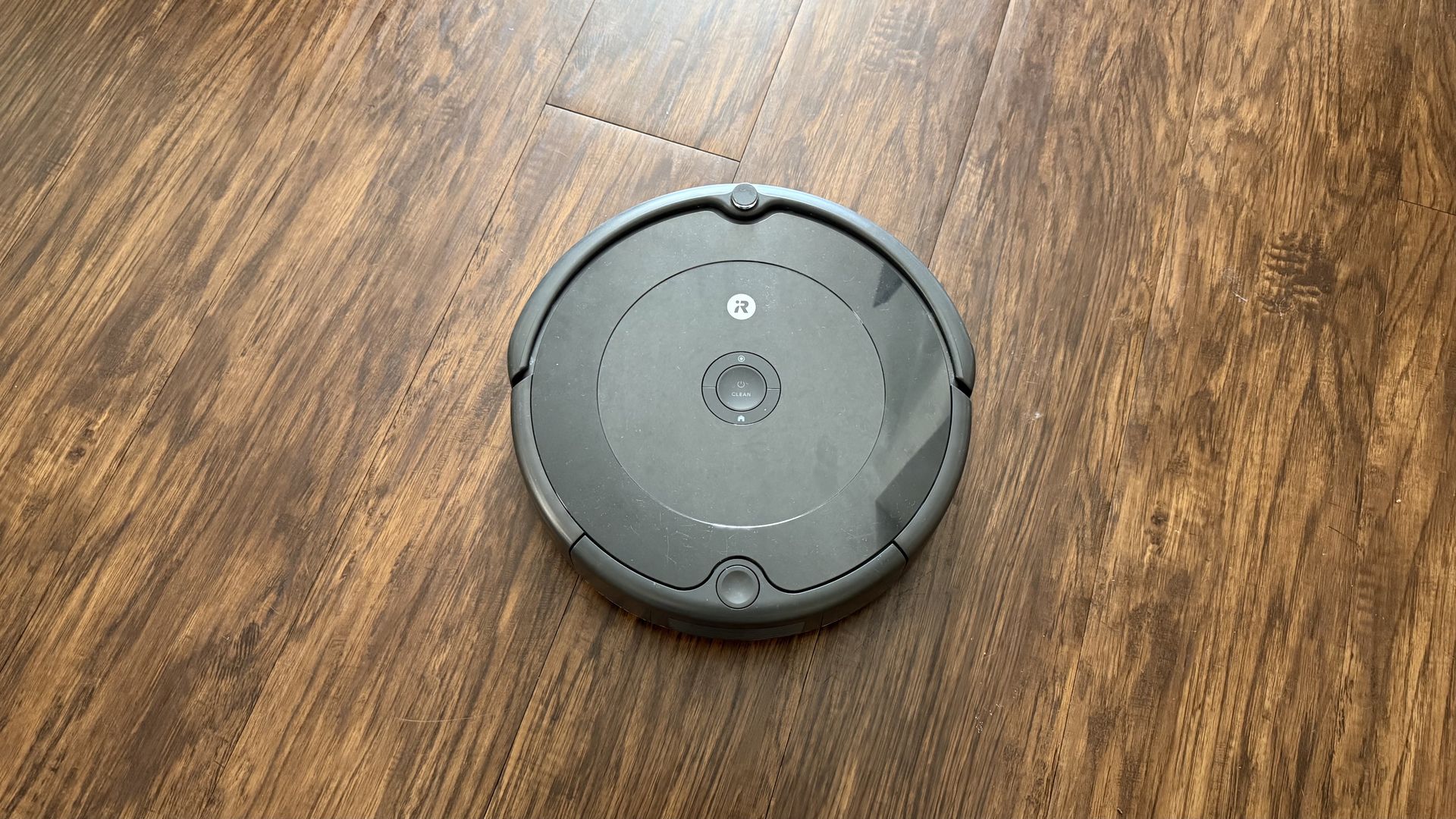 Black robotic vacuum cleaner on a medium-brown wood floor with visible grain patterns, center view from above with 'CLEAN' button visible.