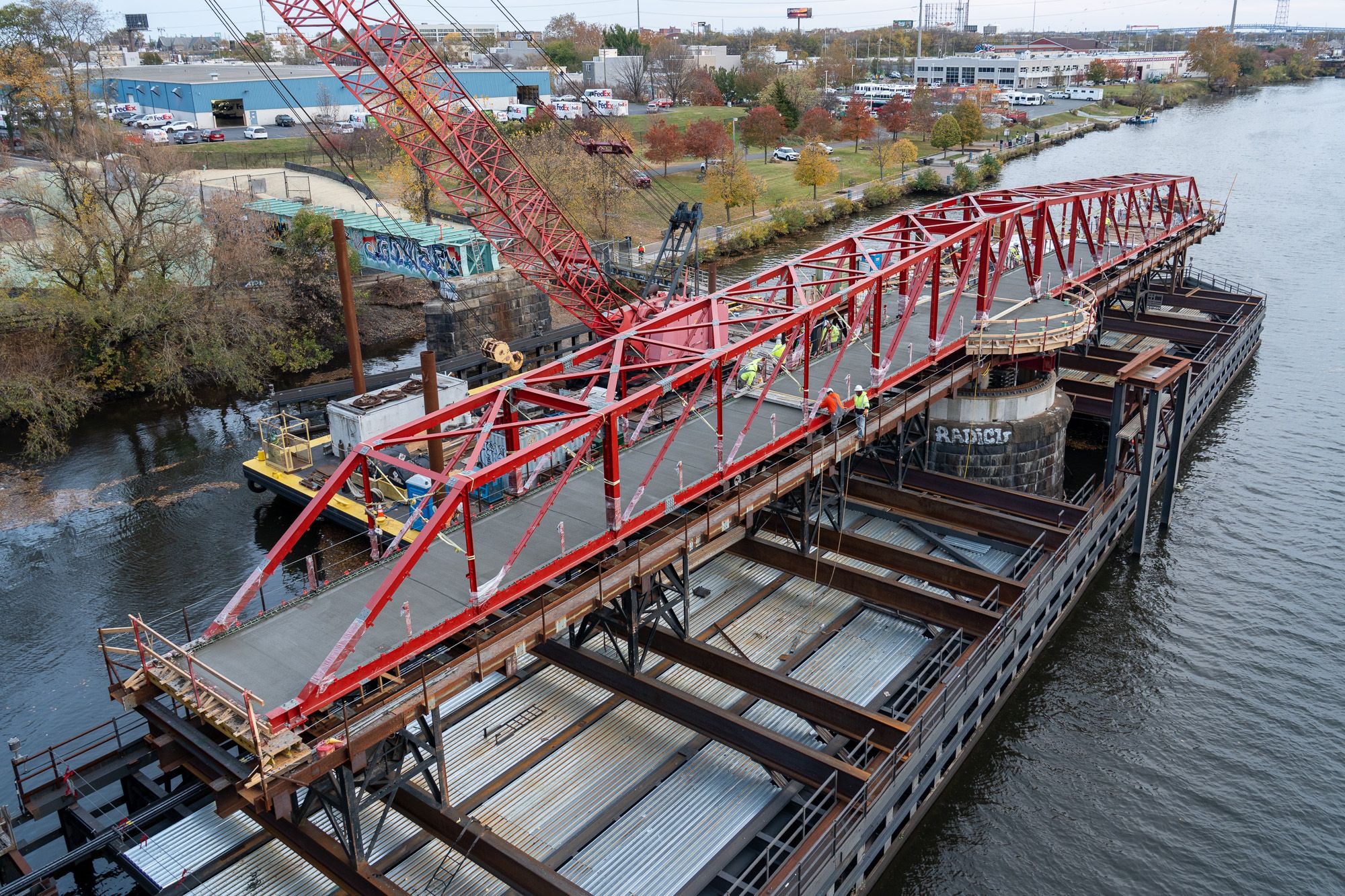 A red truss bridge under construction over a river with workers in high-visibility clothing and a large crane on a barge. Trees and industrial buildings are visible in the background.