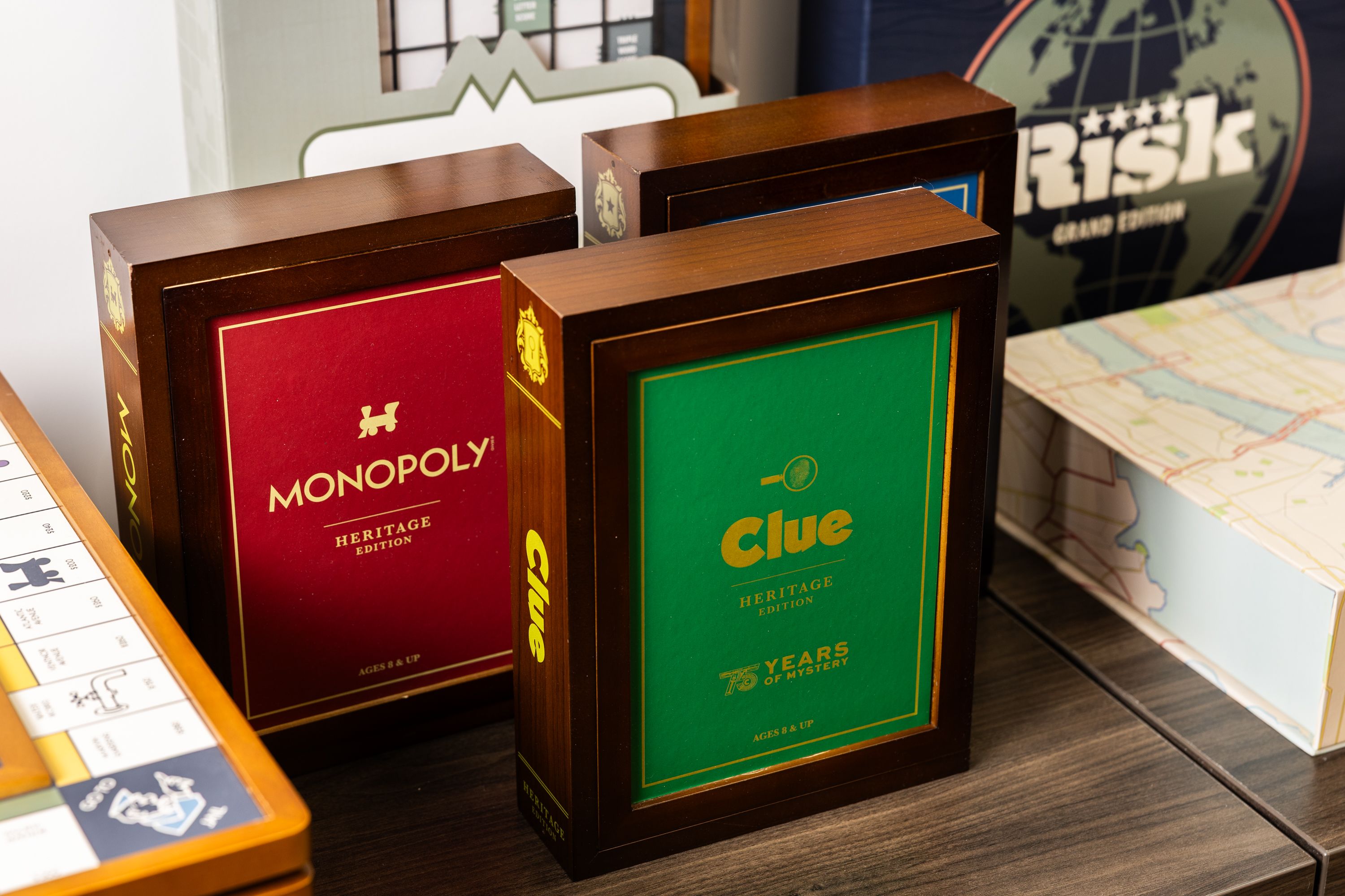 Heritage edition board games Monopoly in red box, Clue in green box celebrating 75 years, and Risk Grand Edition box on a wooden table with part of a Monopoly board visible.