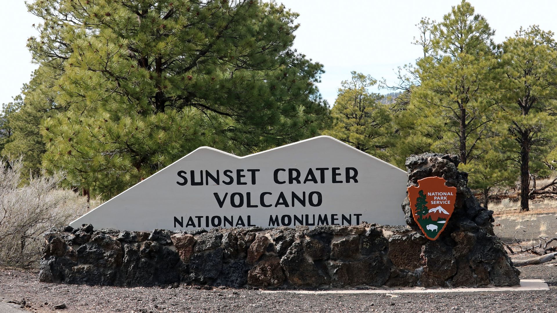 Stone and concrete sign reading "Sunset Crater Volcano National Monument" with National Park Service emblem, surrounded by green pine trees and dry brush under clear sky.
