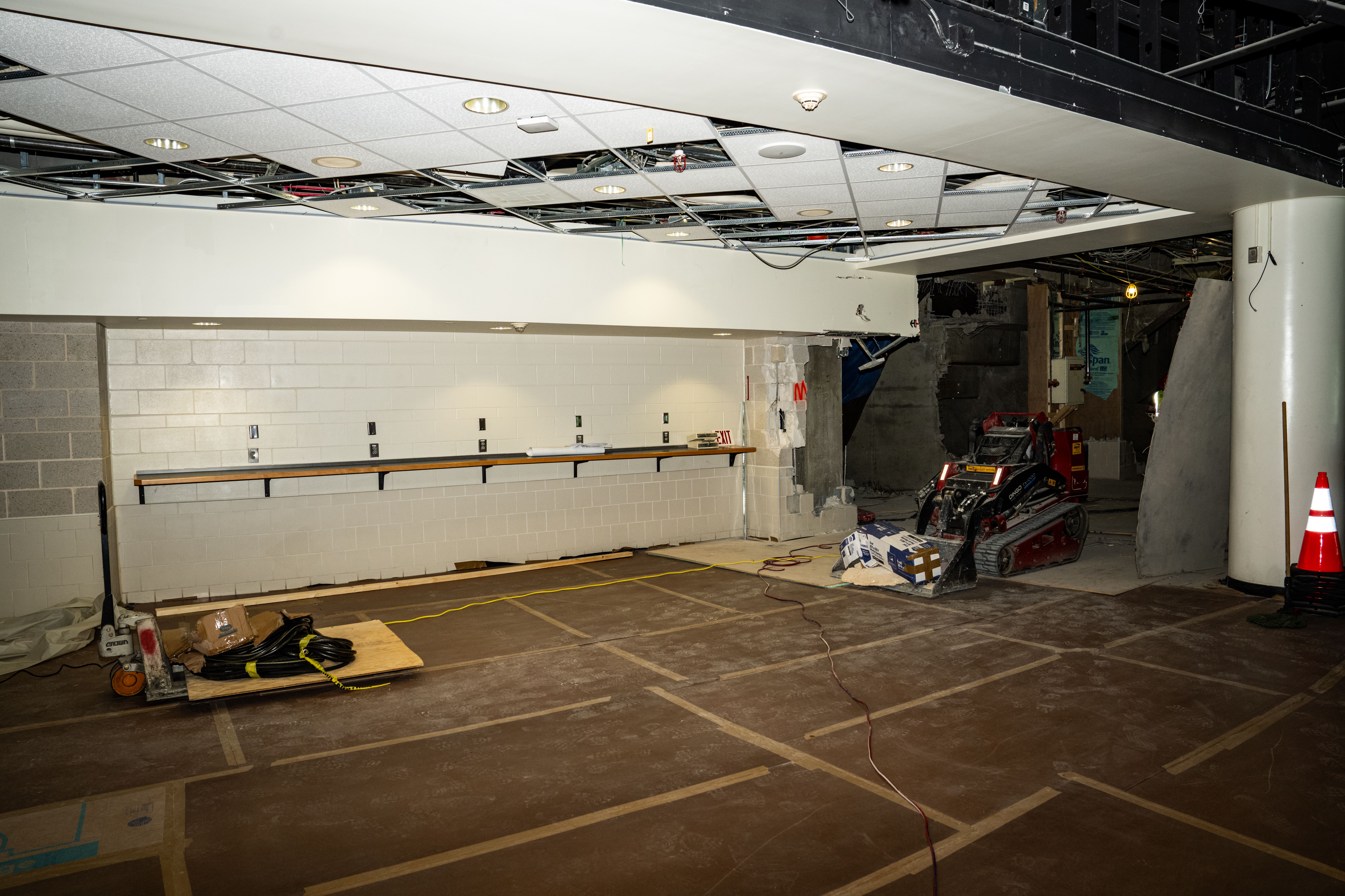 Indoor construction site with damaged ceiling panels, a small red excavator, scattered cables, an orange traffic cone, and partially demolished white and gray walls.