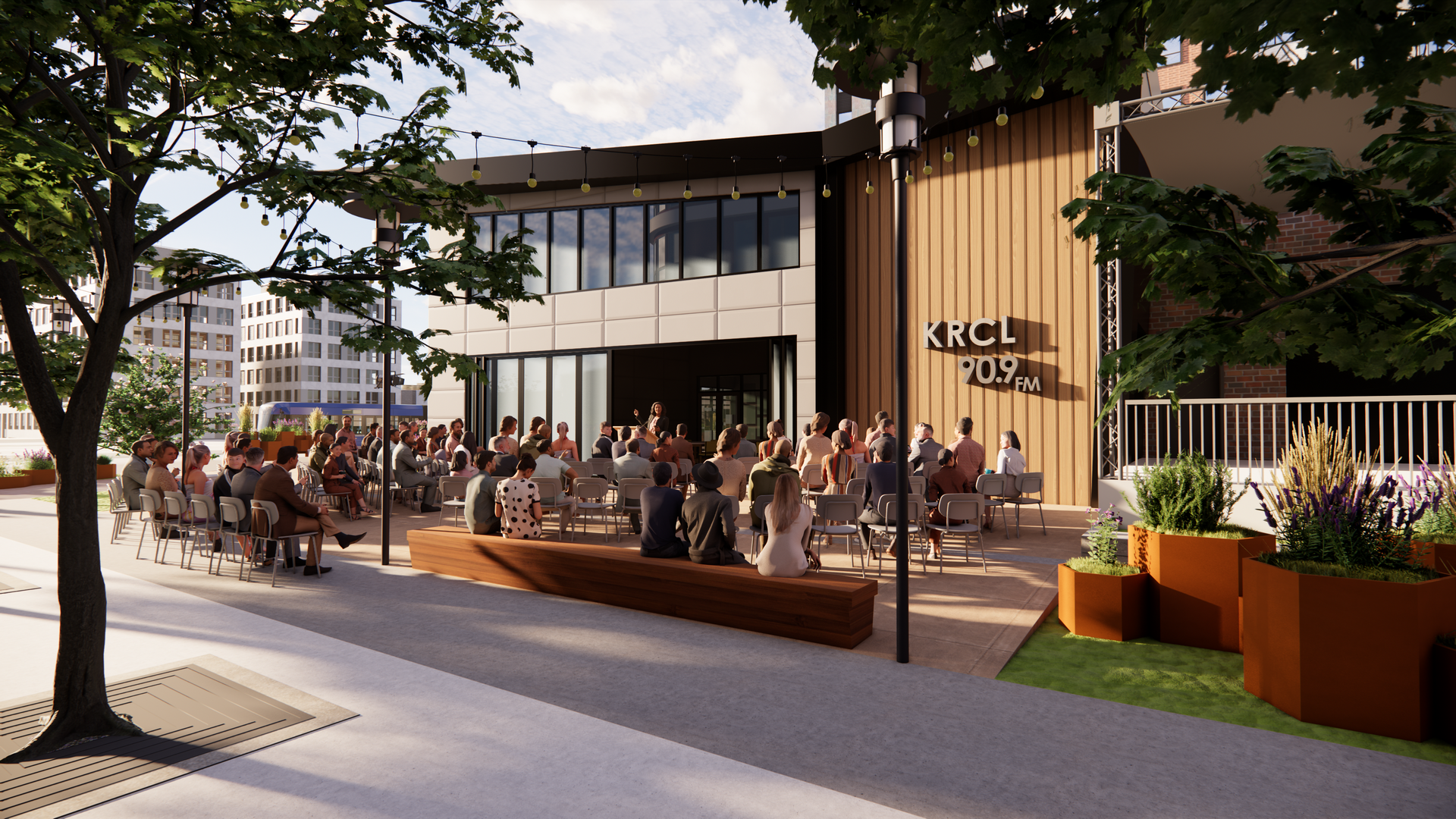 Outdoor city plaza event: a speaker addresses a seated crowd in front of a modern building with "KRCL 90.9 FM". String lights, trees, and orange planters frame the scene.