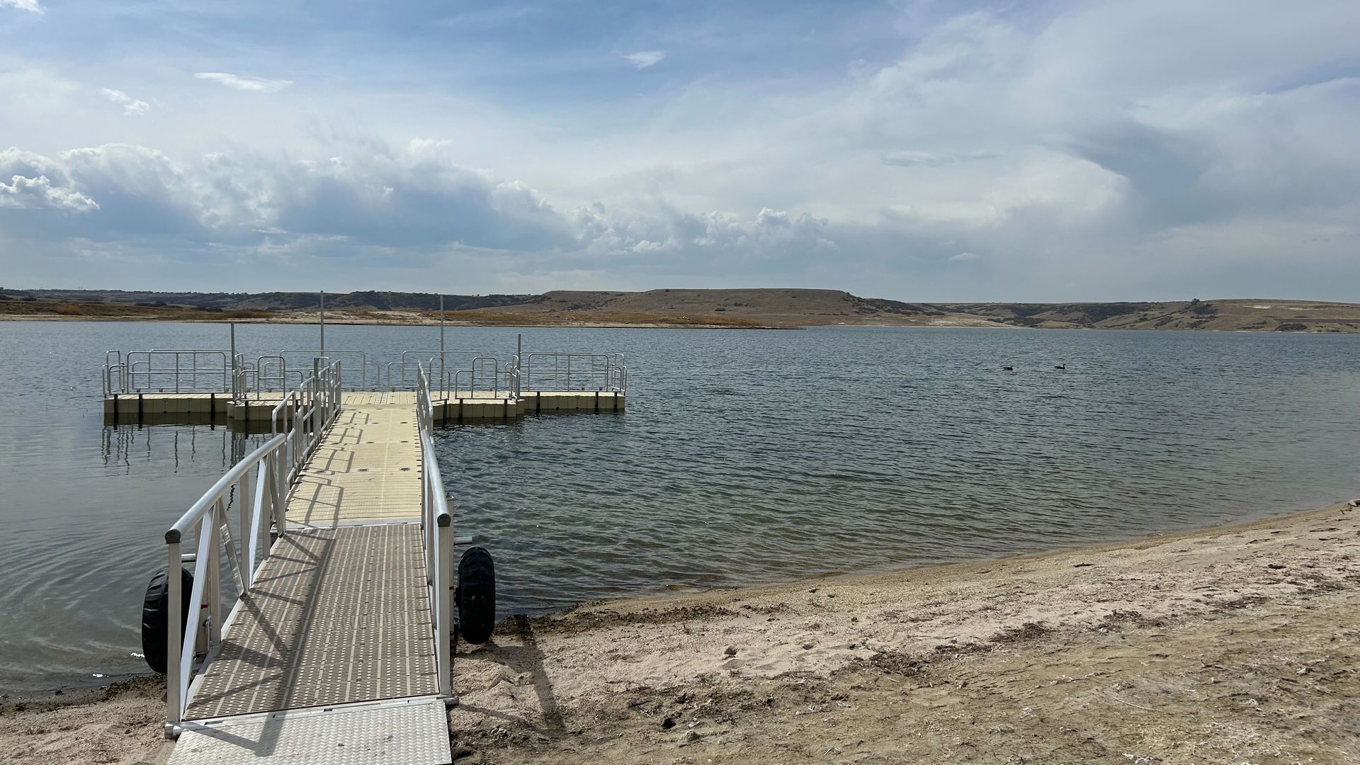 Metal dock with railing and floating platforms extends from a sandy shore into a calm lake; a black tire rests on the pier. Distant barren hills beneath a blue, partly cloudy sky.