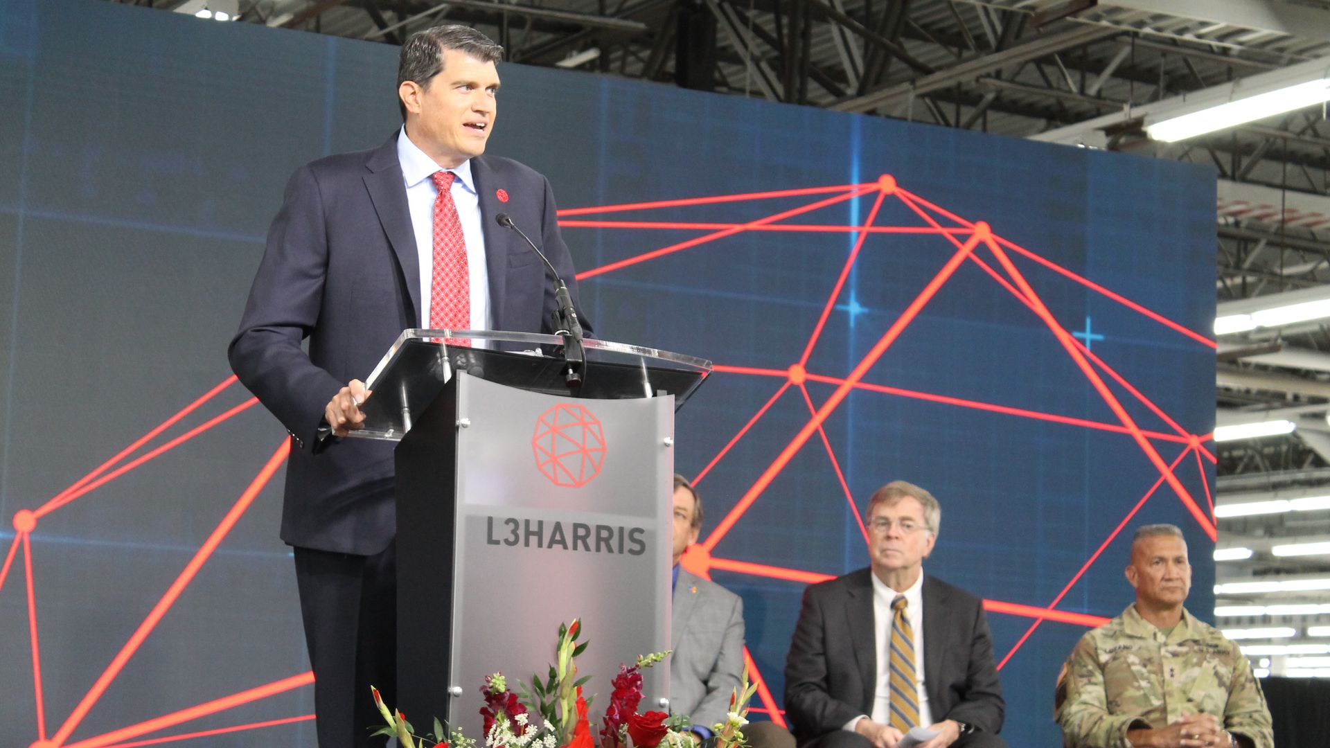 Man in dark suit and red tie speaking at an L3Harris podium with a red and blue geometric digital network graphic backdrop, two seated men in suit and military uniform behind him.