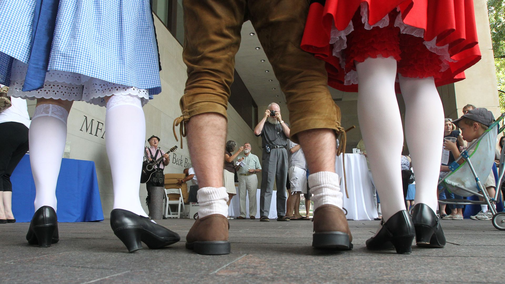 Low-angle view of legs in white tights and colorful skirts (blue gingham and bright red with lace) lining a stage; in the background, a photographer, attendees, and a guitarist perform.