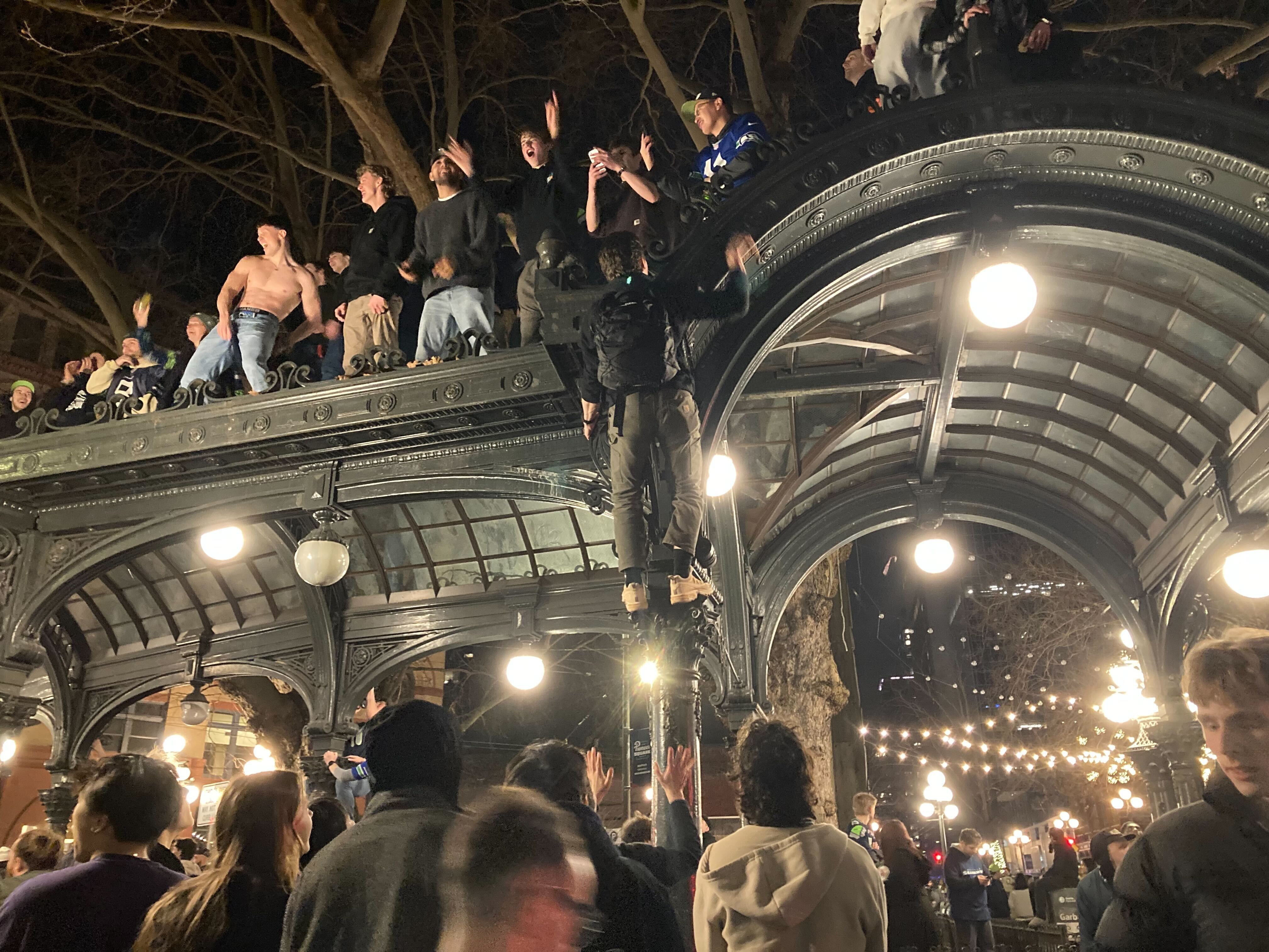 Nighttime outdoor scene with a crowd gathered under and on a decorative metal and glass pavilion. People are standing, some climbing the structure, with bare trees and hanging lights visible.