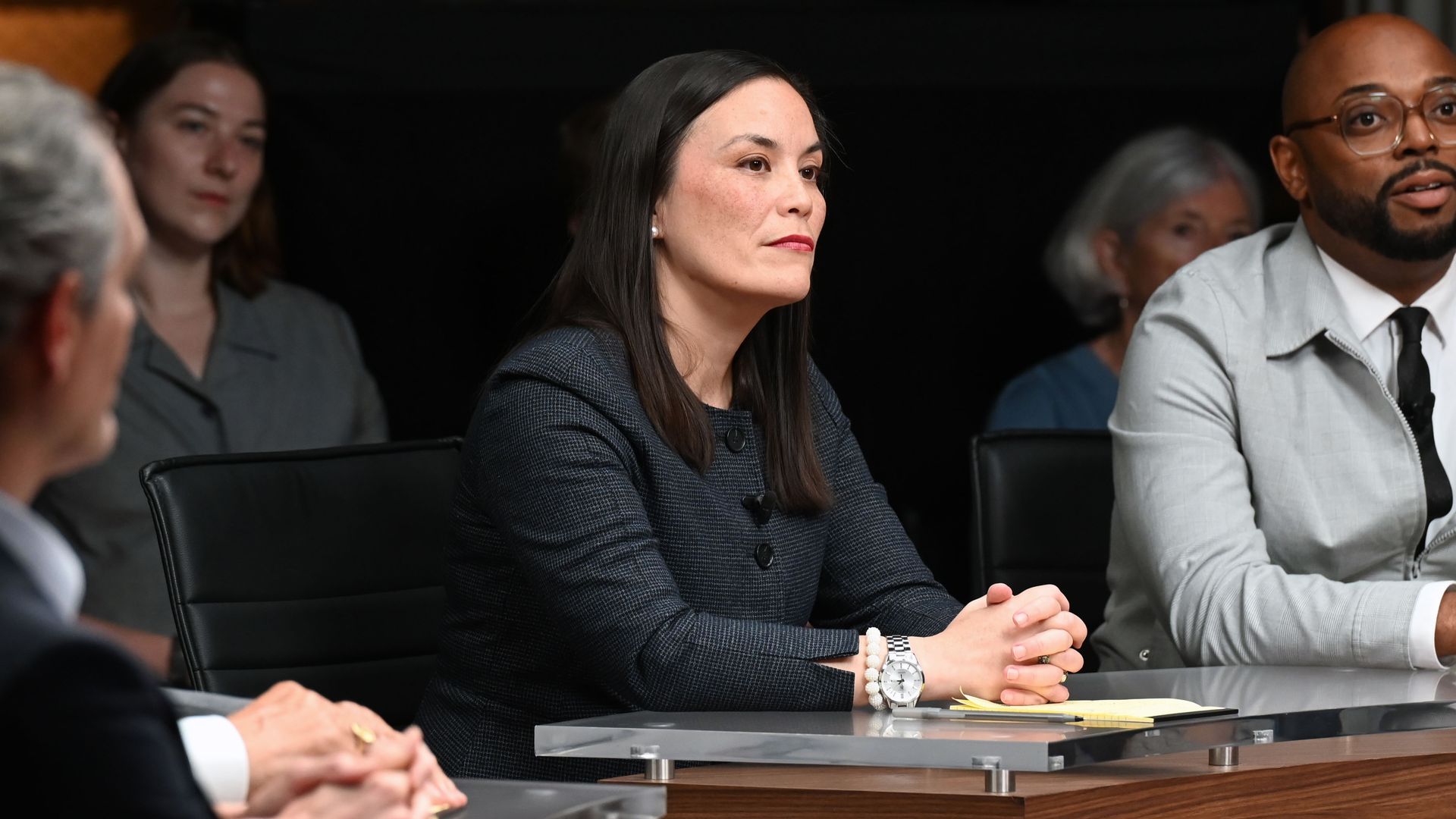 San Antonio Mayor Gina Ortiz Jones in an episode of "Breaking the Deadlock" on PBS. Jones is seen with her hands clasped together on a conference table; she is wearing a dark-colored blazer as she listens.