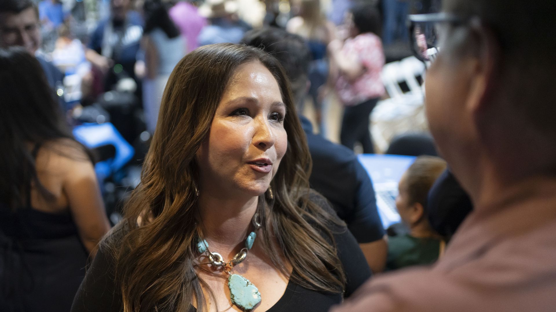 Adelita Grijalva, with long brown hair and wearing a large turquoise necklace and black top talking to a man in glasses, with numerous people in the background. 