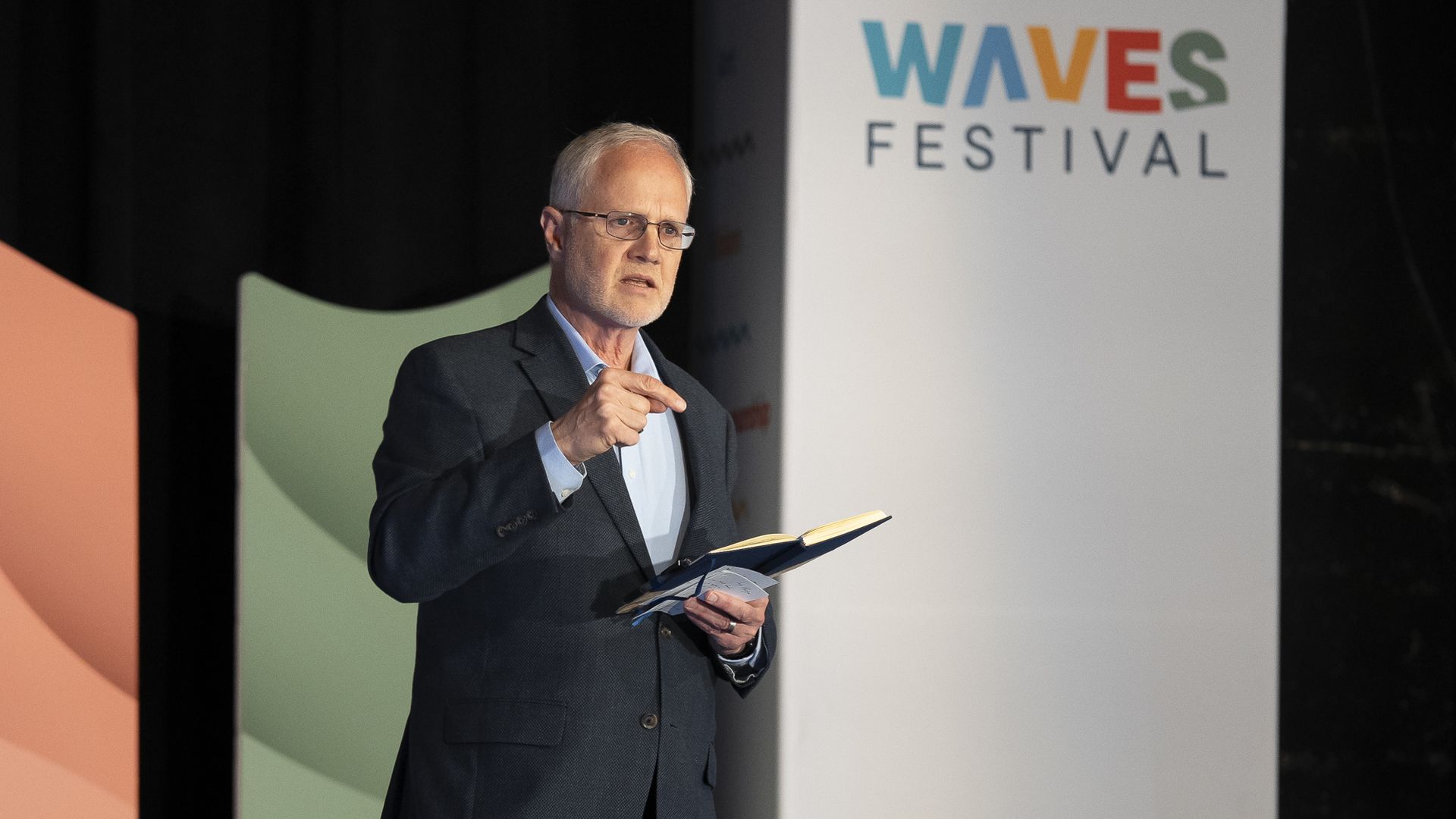 Grant Oliphant standing in front of a colored background and a sign that reads "Waves Festival" in a rainbow