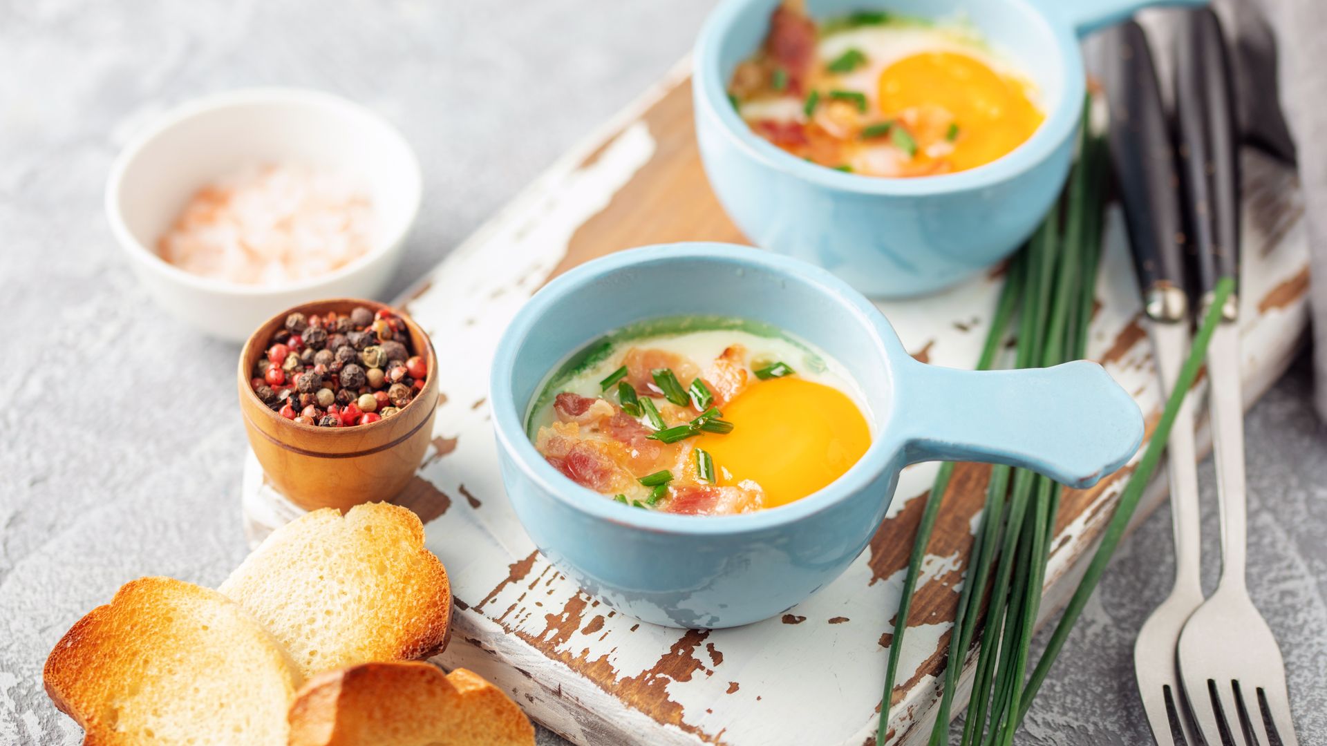 Two light-blue ramekins hold Oeufs cocotte with gruyere, bacon and chives on a rustic white board.