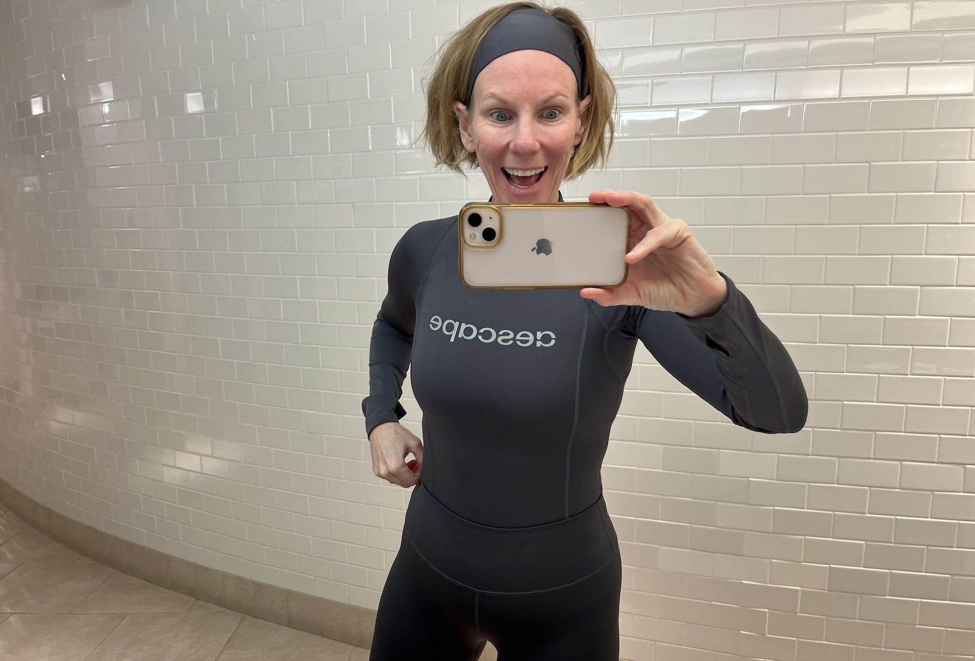 Carrie Shepherd in a gray wetsuit taking a selfie in front of white tiled wall, holding phone with a clear case, looking excited and smiling.