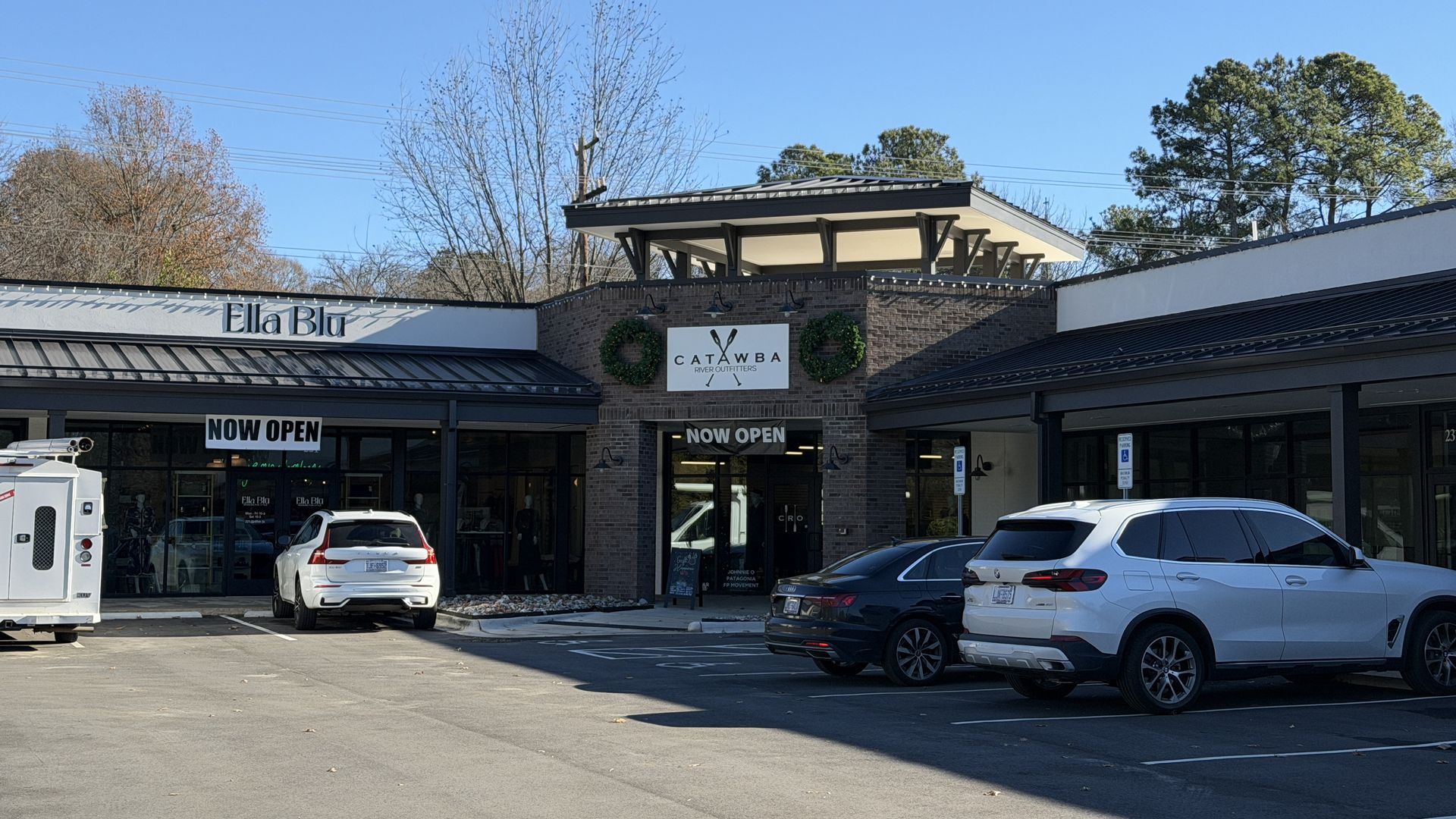 Outdoor view of a shopping plaza with stores Ella Blu and Catawba River Outfitters, both displaying 'NOW OPEN' signs, parked cars, clear blue sky, and leafless trees in background.