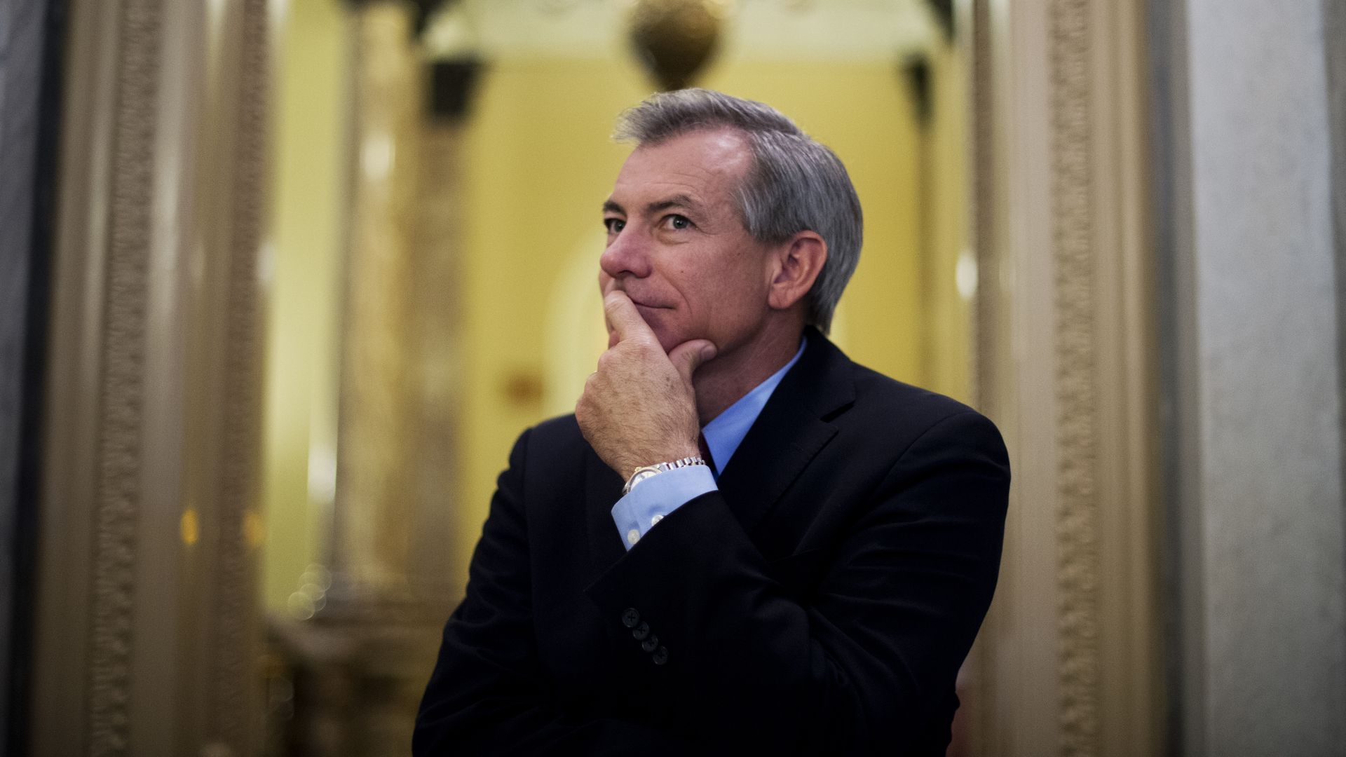 David Schweikert in a dark suit and blue shirt, resting his chin on his hand, standing in an elegant hallway with ornate columns and warm lighting.