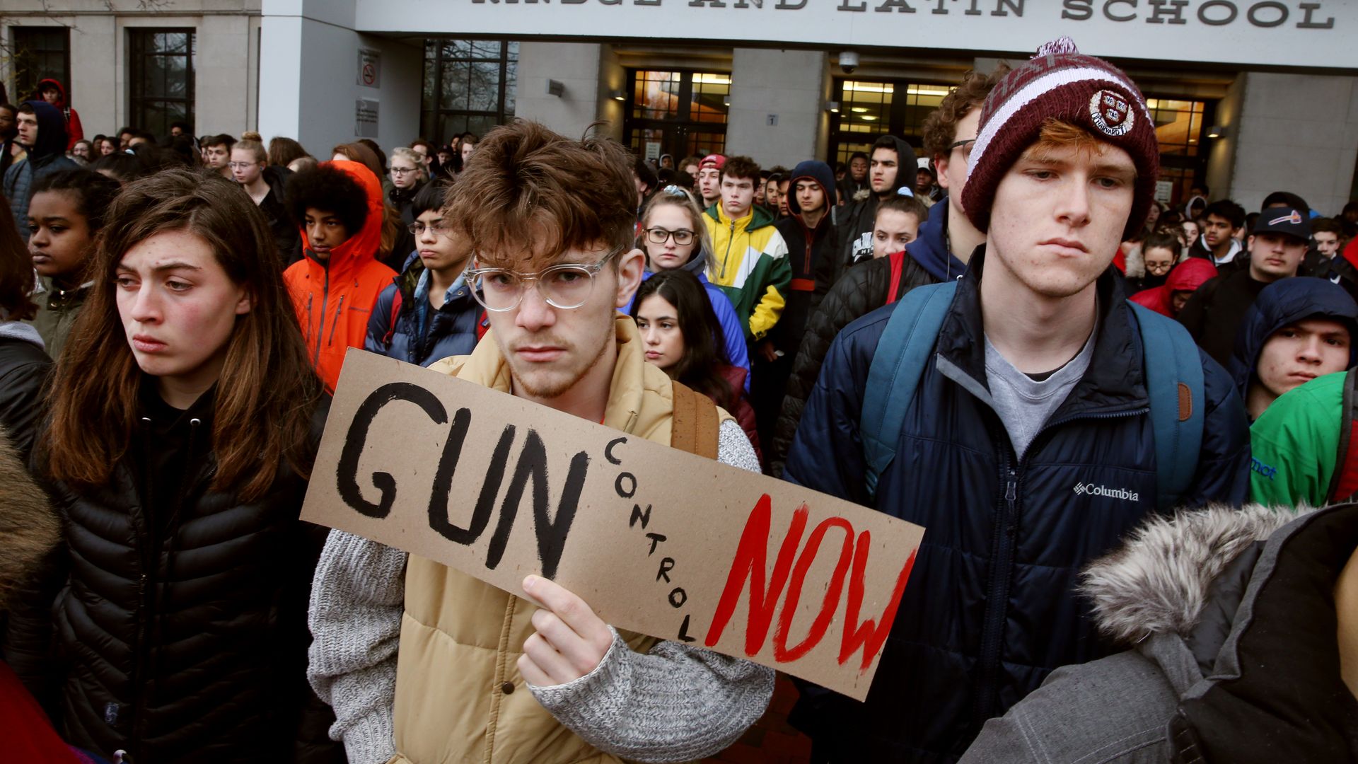 protestors outside of a school hold a sign that says gun control now