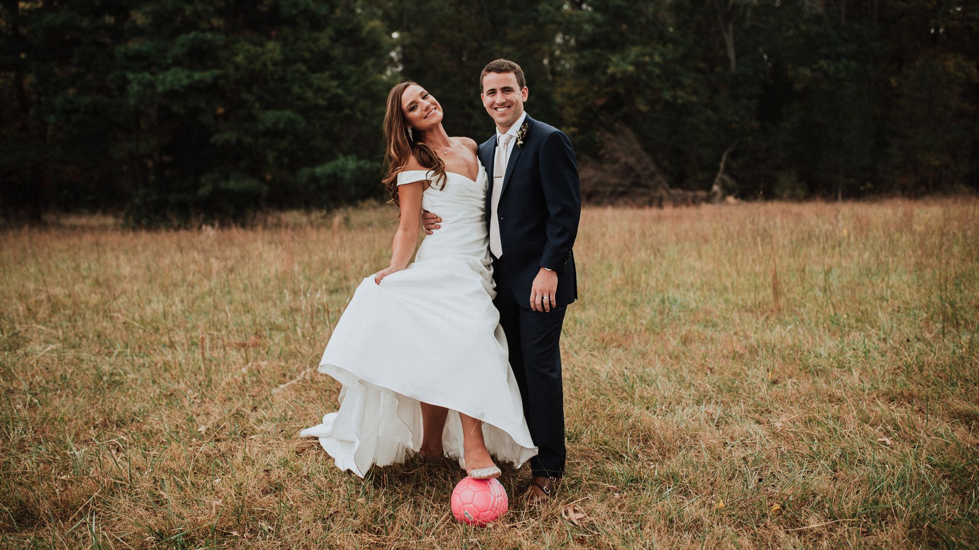Two people, one in a wedding dress and the other in a tuxedo, posing over a soccer ball 