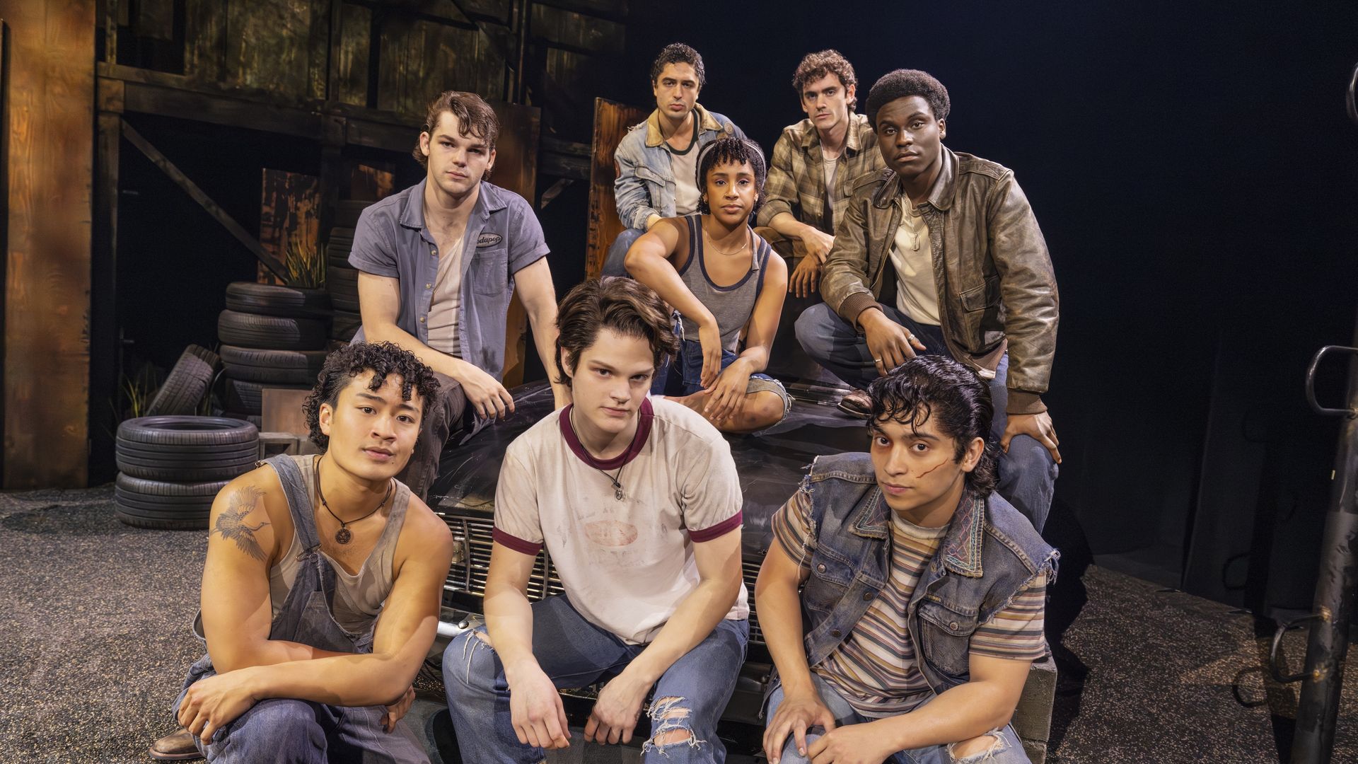 A diverse group of young actors sits on a stage set with tires and weathered wood, wearing denim jackets and casual tops, posing for the camera against a dark backdrop.