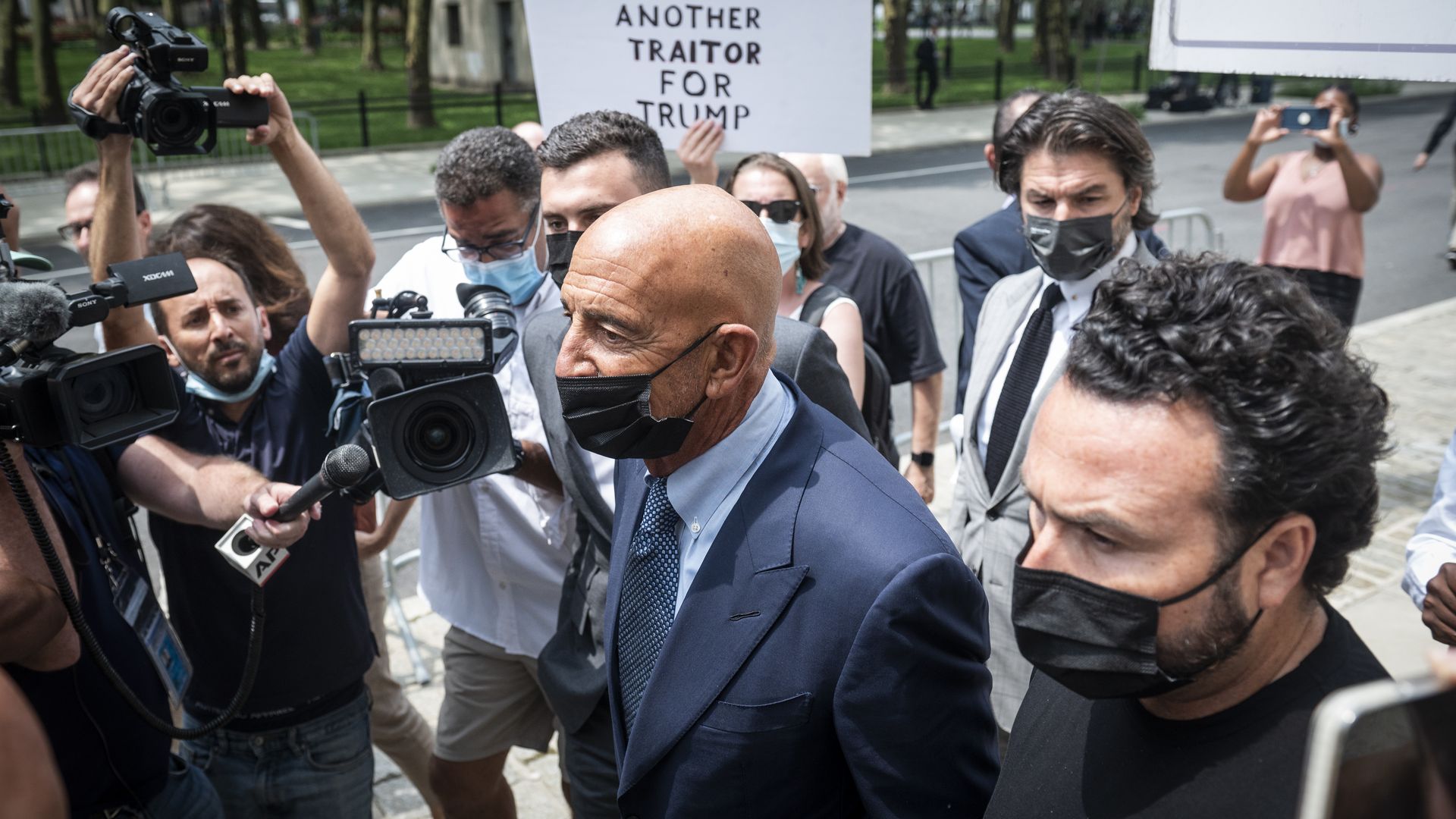 Photo of a masked Tom Barrack walking through a crowd of people and reporters