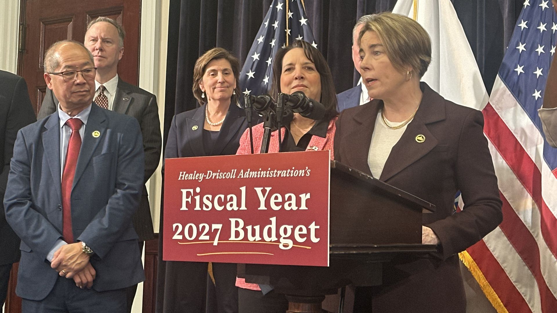 Gov. Maura Healey stands at a lectern, surrounded by cabinet members and other state leaders, as she unveils the fiscal 2027 budget proposal at the Massachusetts State House. 