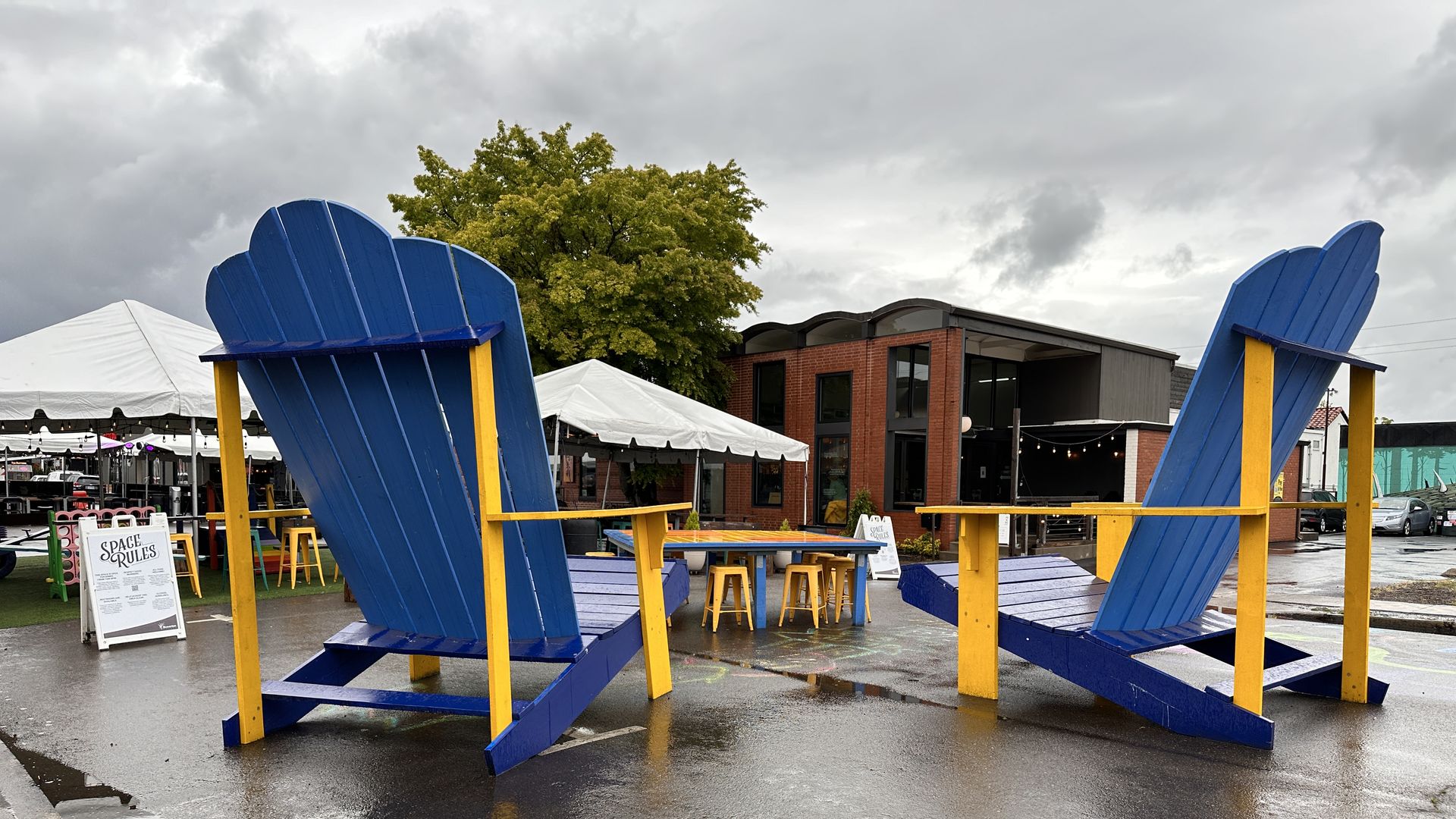 Two large blue and yellow beach chairs in a wet plaza in front of white rain covers and a brick building and tree