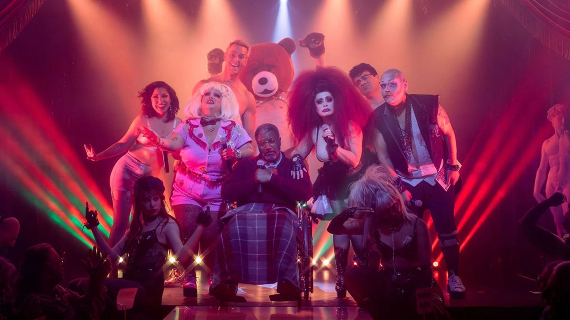 A diverse group of performers in colorful, eclectic costumes on stage with red curtains and bright spotlights, including a person in a bear costume and one in a wheelchair.