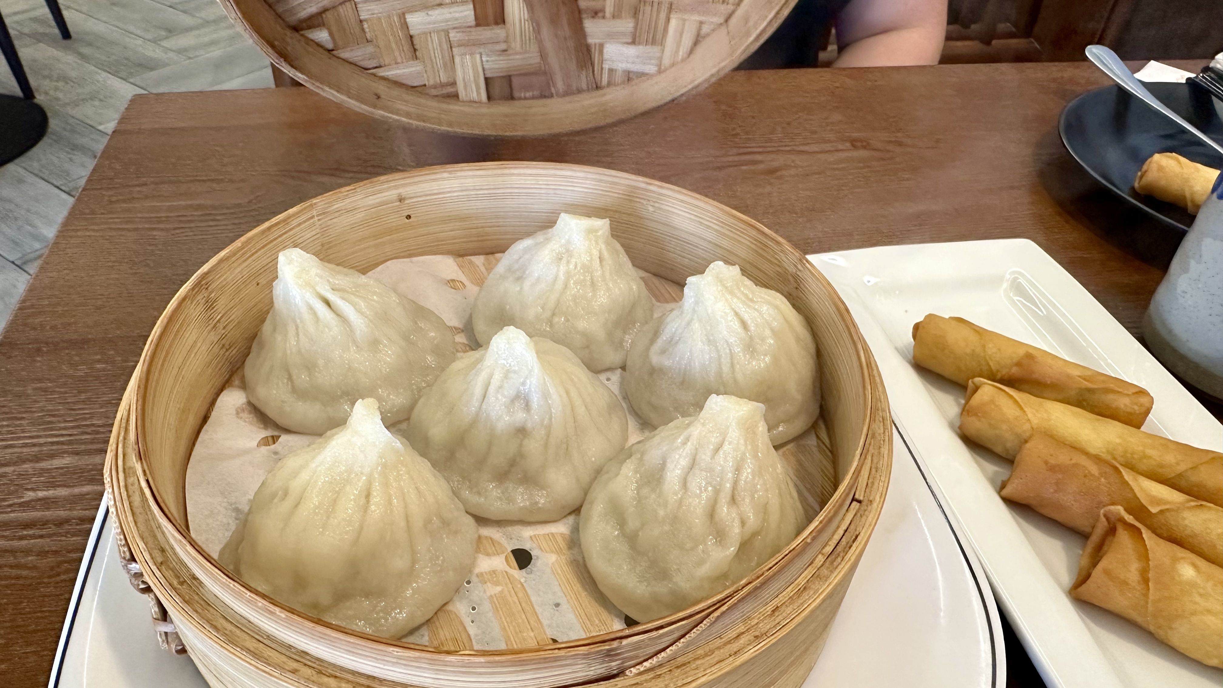 A round bamboo steamer basket holds plump steamed dumplings on parchment, sitting on a wooden table beside a plate of golden spring rolls.
