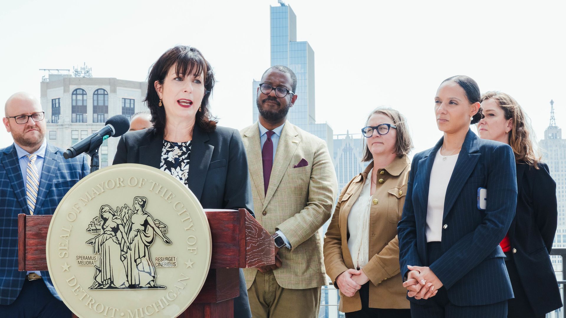 Stella speaks at a podium with Detroit's seal, with other officials and the city skyline behind her