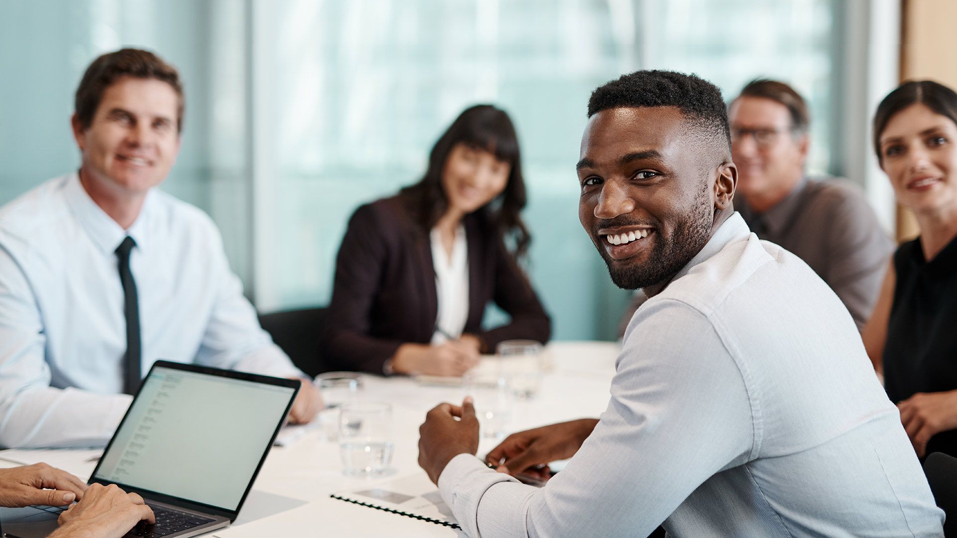 Smiling professional in a business meeting, seated at a conference table with colleagues and a laptop.