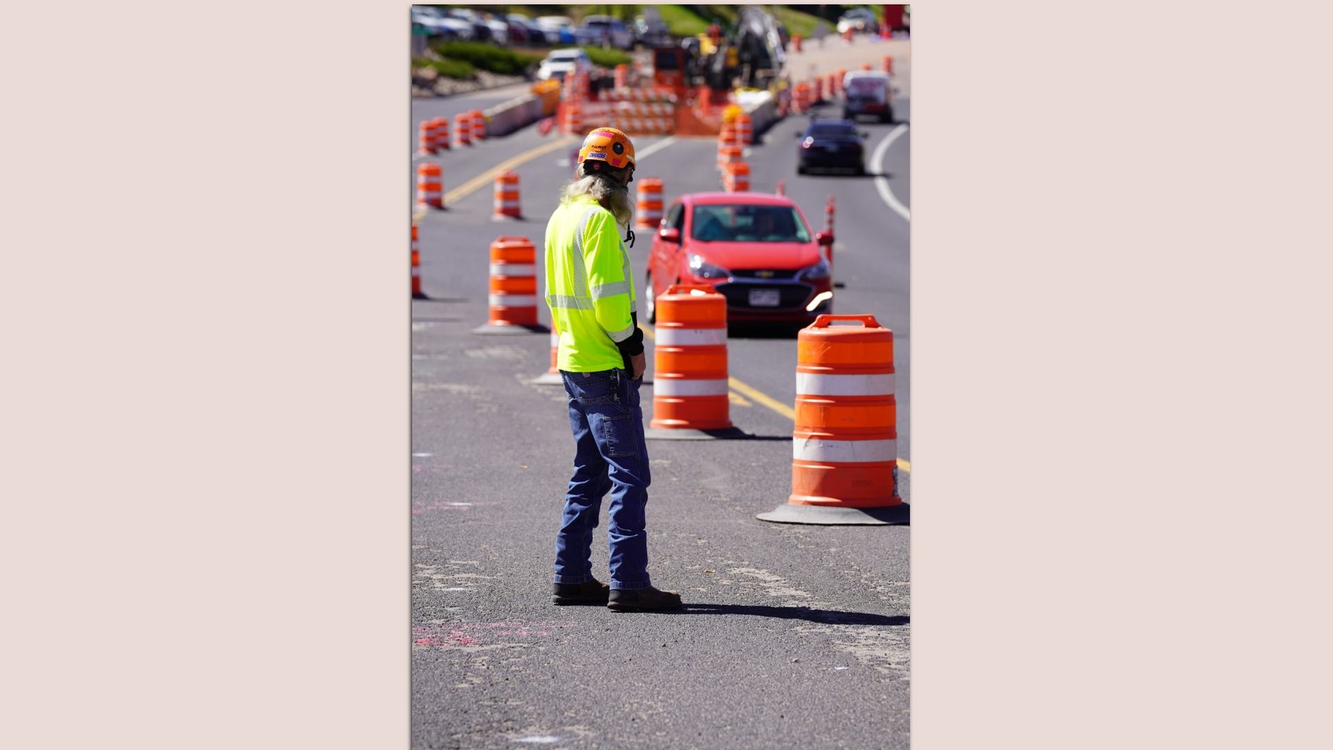 A road worker in a high-visibility vest stands on a road between traffic cones.