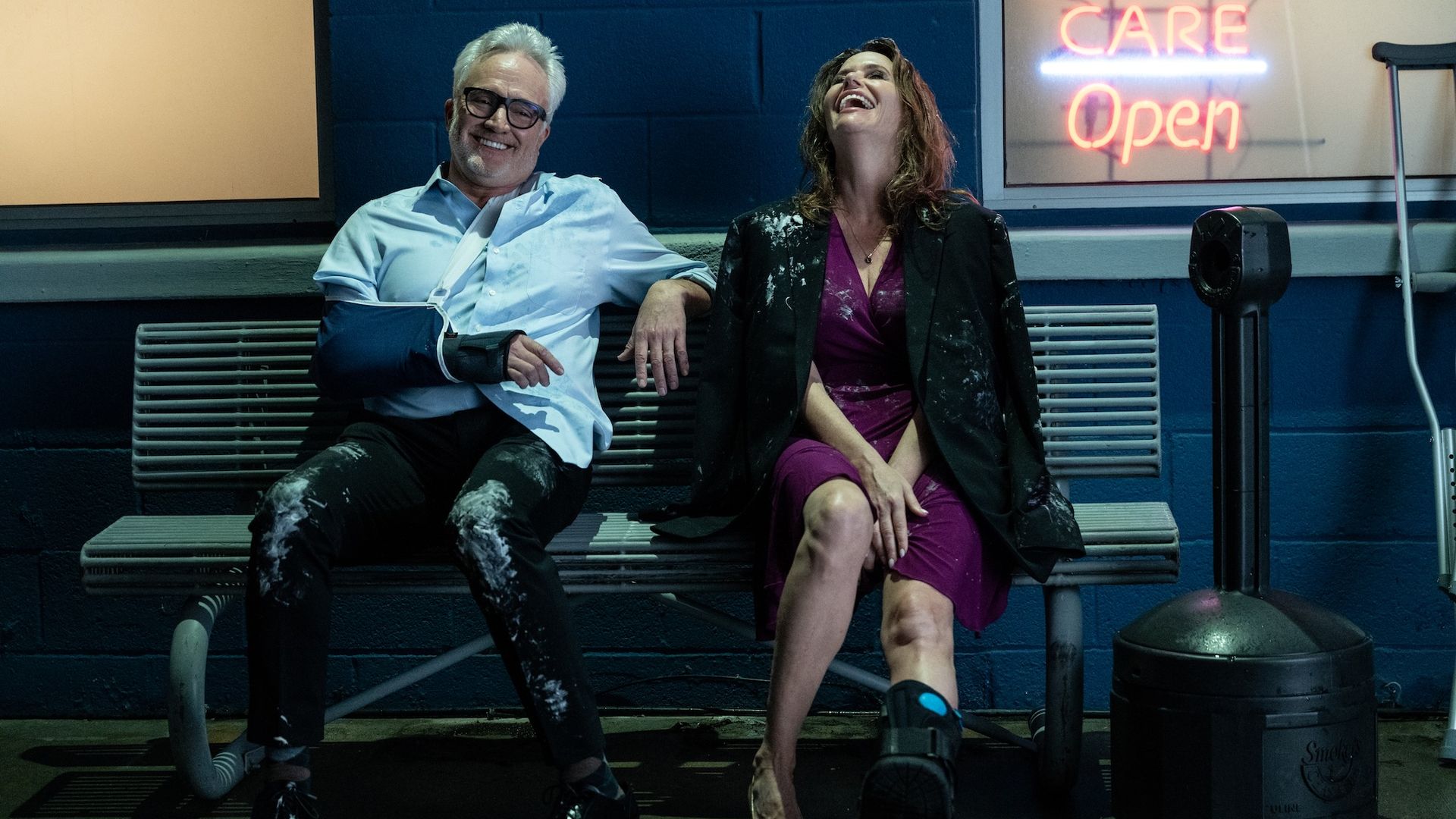 Bradley Whitford with a cast and sling on his left arm and Amy Landecker with a knee brace and crutches sit on a bench outside an urgent care, both laughing with paint splattered on their clothes.