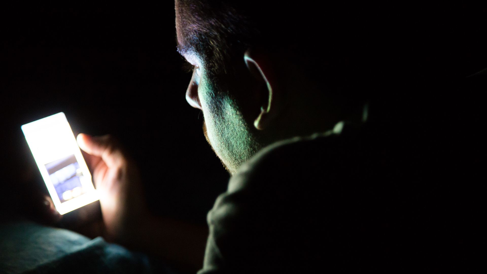 A mani in the dark looks at a smartphone, which illuminates his face