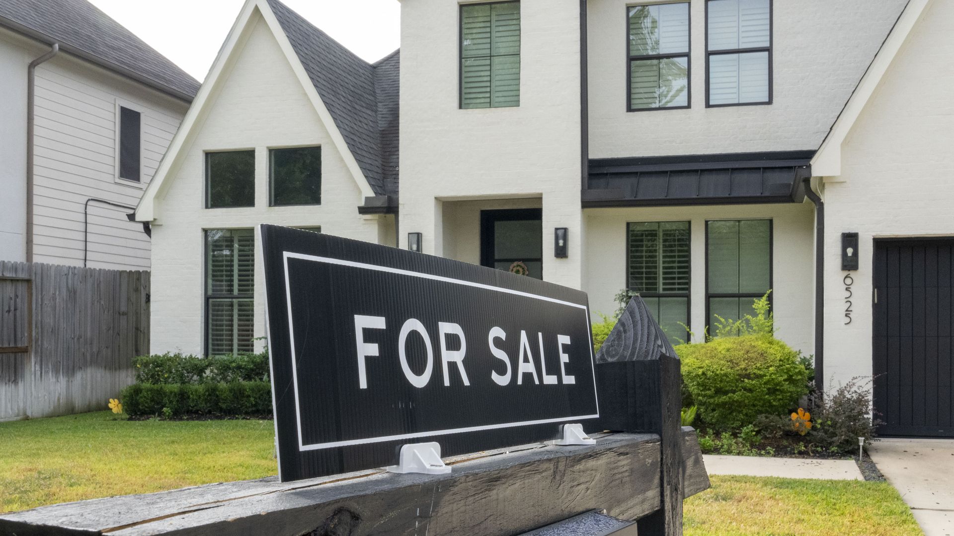 A "FOR SALE" sign on dark wooden post in front of a white two-story house with green bushes, lawn, and black-framed windows.