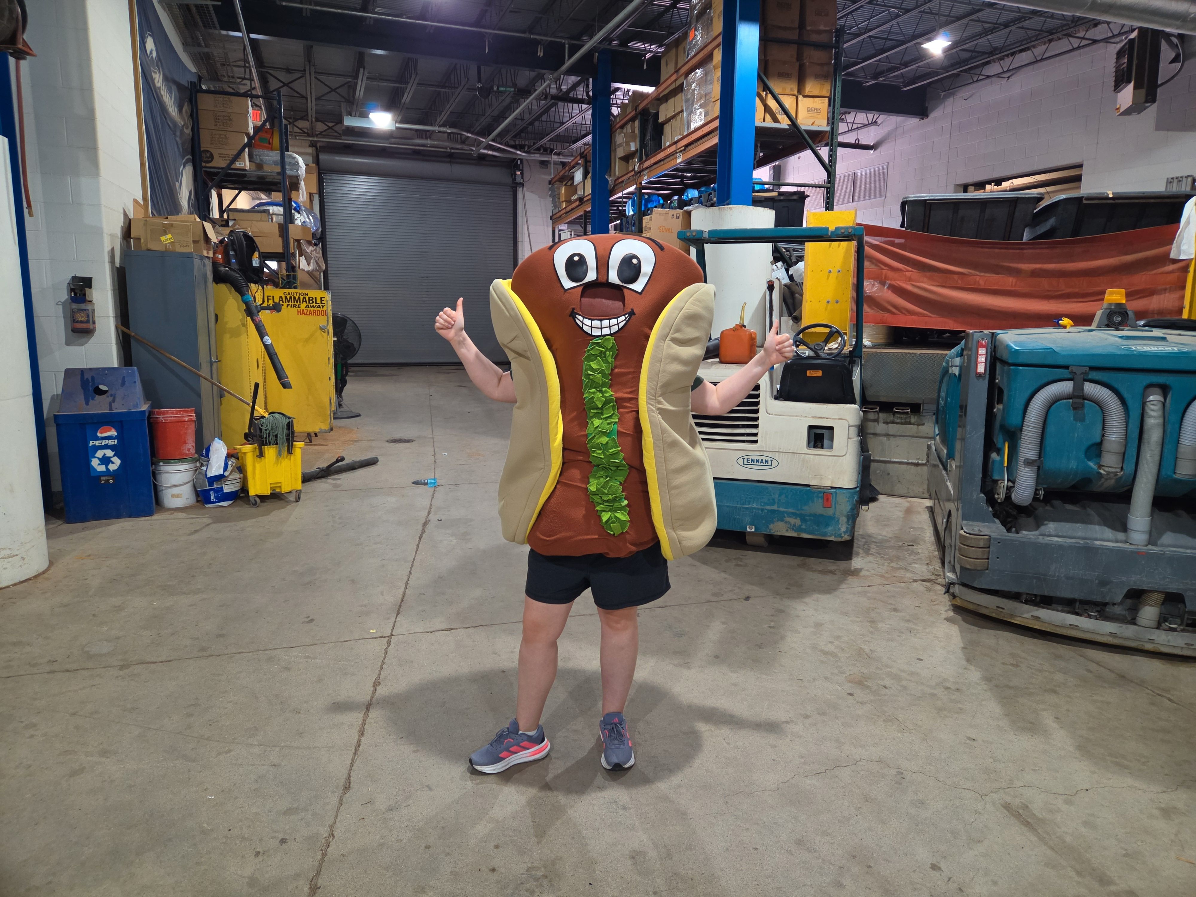 Alissa in a cartoon hot dog costume with relish on it, smiling and giving thumbs up in a warehouse with tall shelves, boxes, a yellow flammable cabinet, and blue industrial machinery.