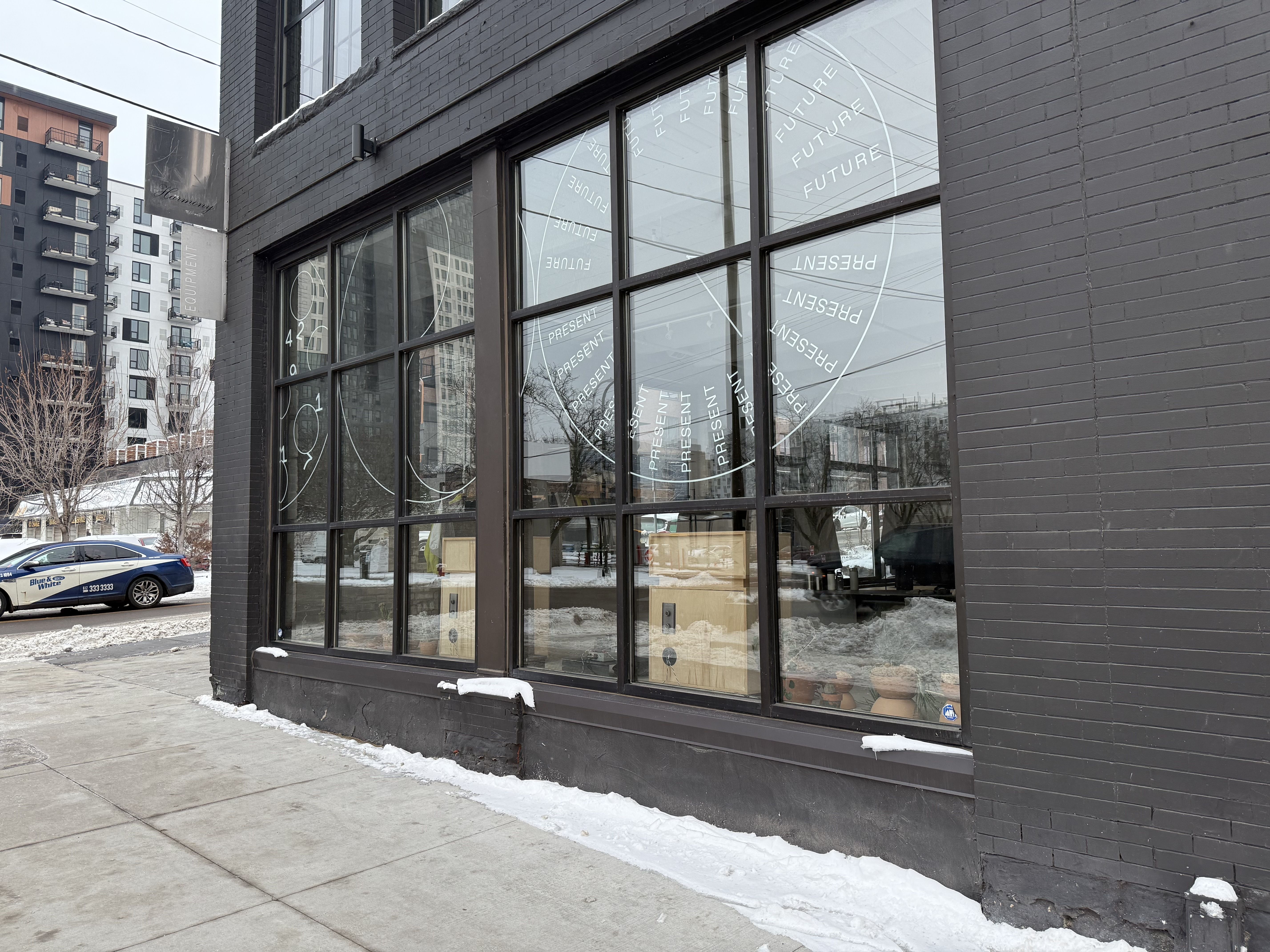 Black brick building with large windows featuring white circular designs and words "PRESENT" and "FUTURE" repeatedly. Snow lines the sidewalk outside and a blue taxi is parked nearby.
