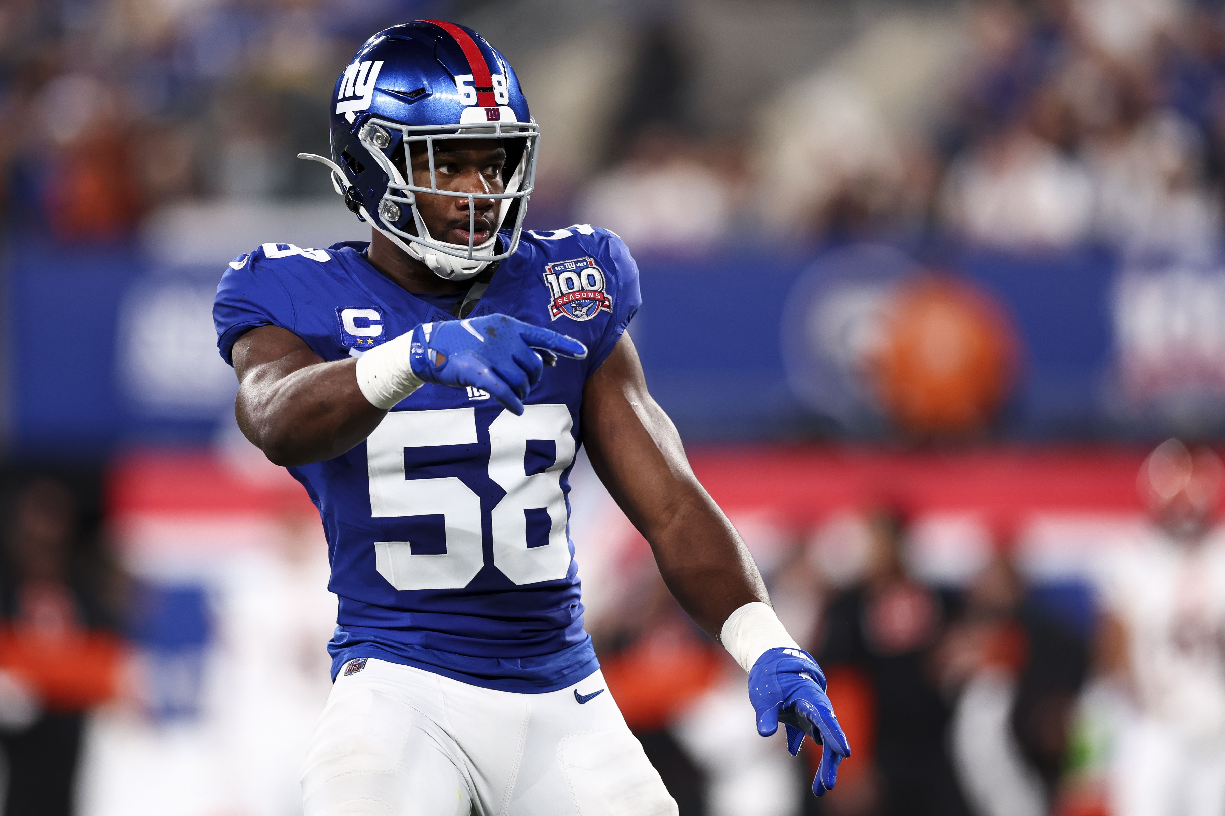 Football player wearing blue jersey number 58 and silver helmet with red stripe, pointing with his left hand during a game, with blurred crowd in the background.