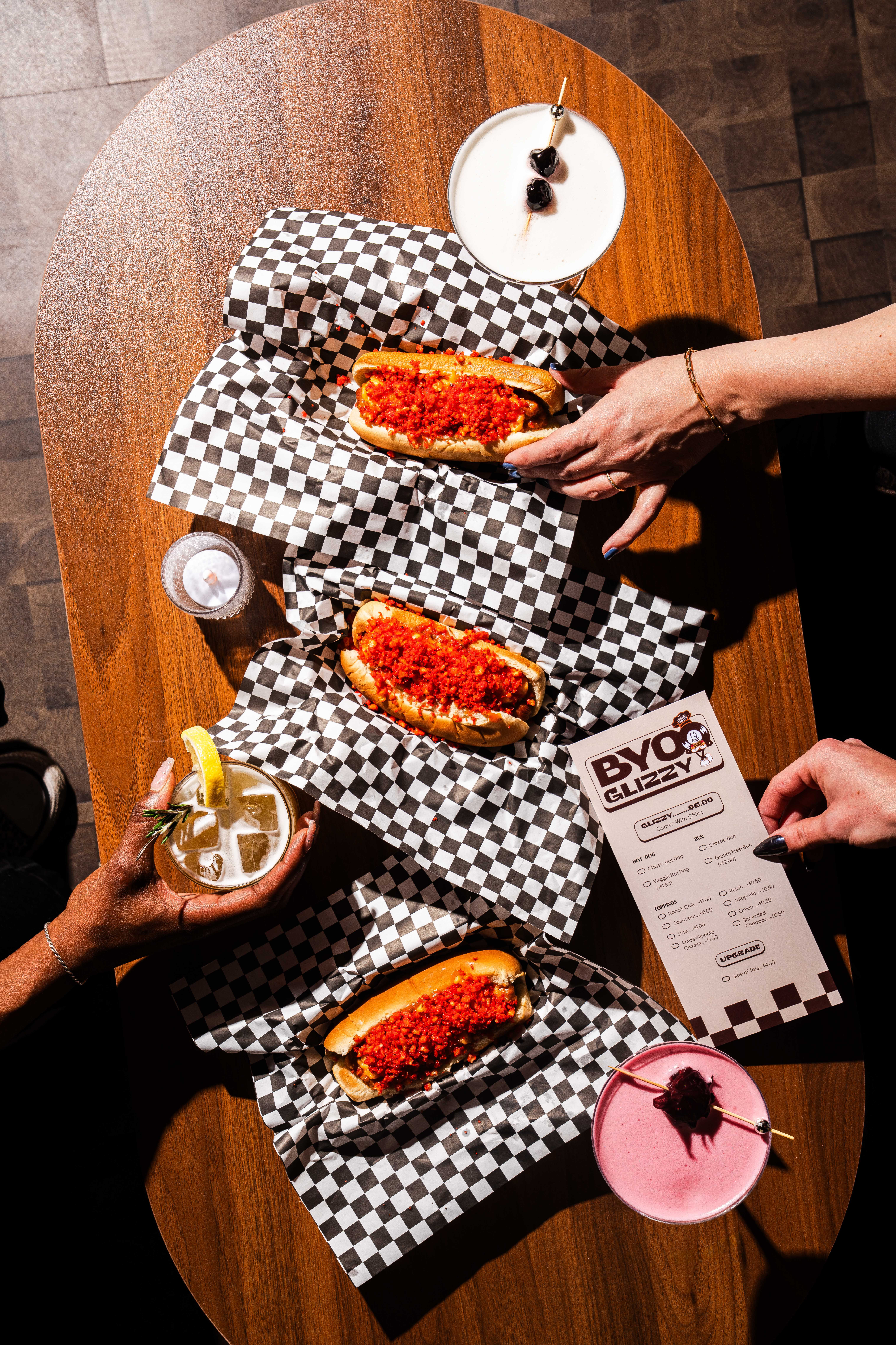 Three chili-topped hot dogs on checkered paper on a wooden table, three cocktails, and two hands holding a hot dog and a BYO Glizzy menu respectively.