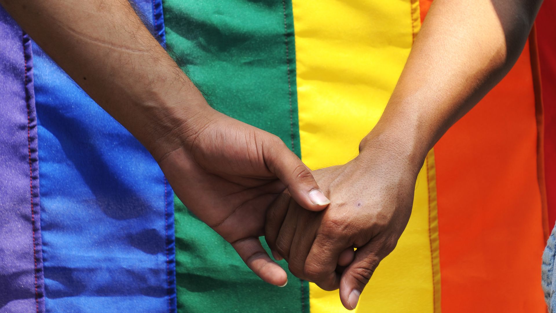 Two people hold hands in front of a pride flag