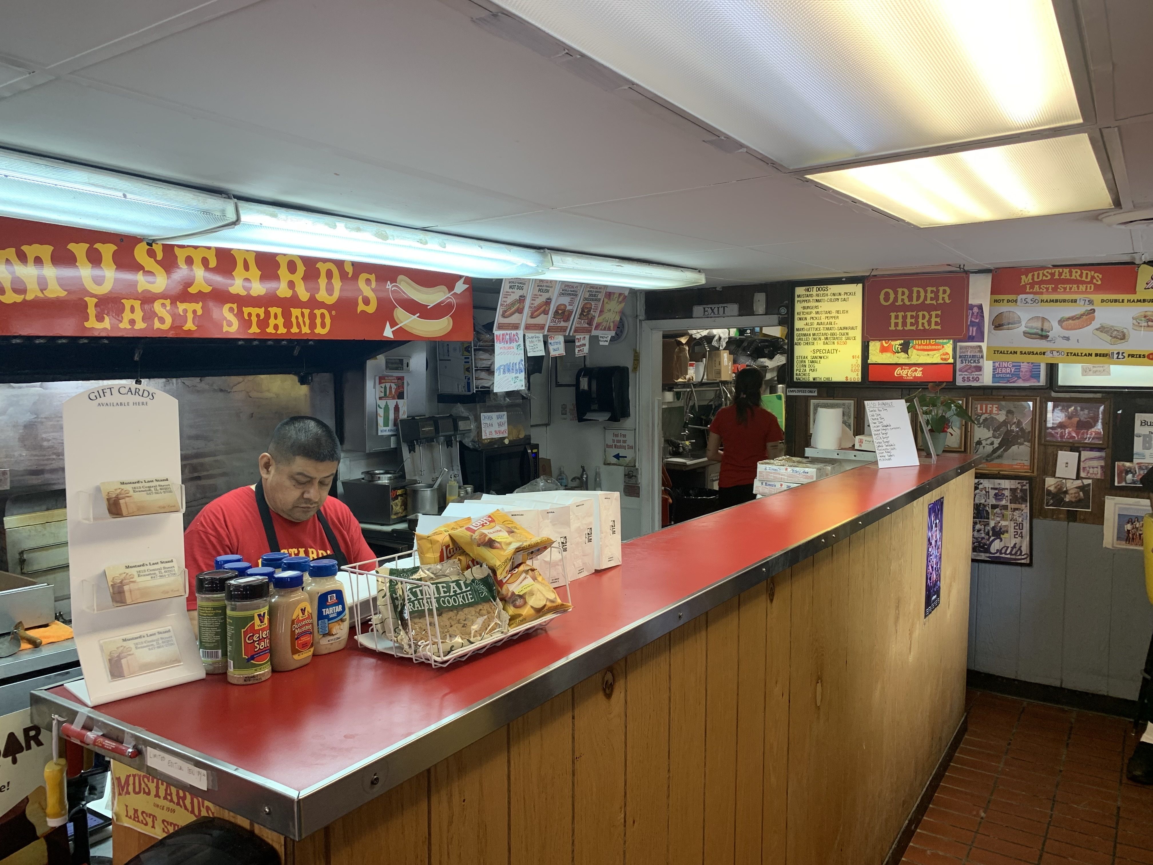 Interior of a fast-food counter with a long red countertop, wooden panels, bright lights, a worker in red, mustard bottles, and a sign reading "Mustard's Last Stand" beside menus.