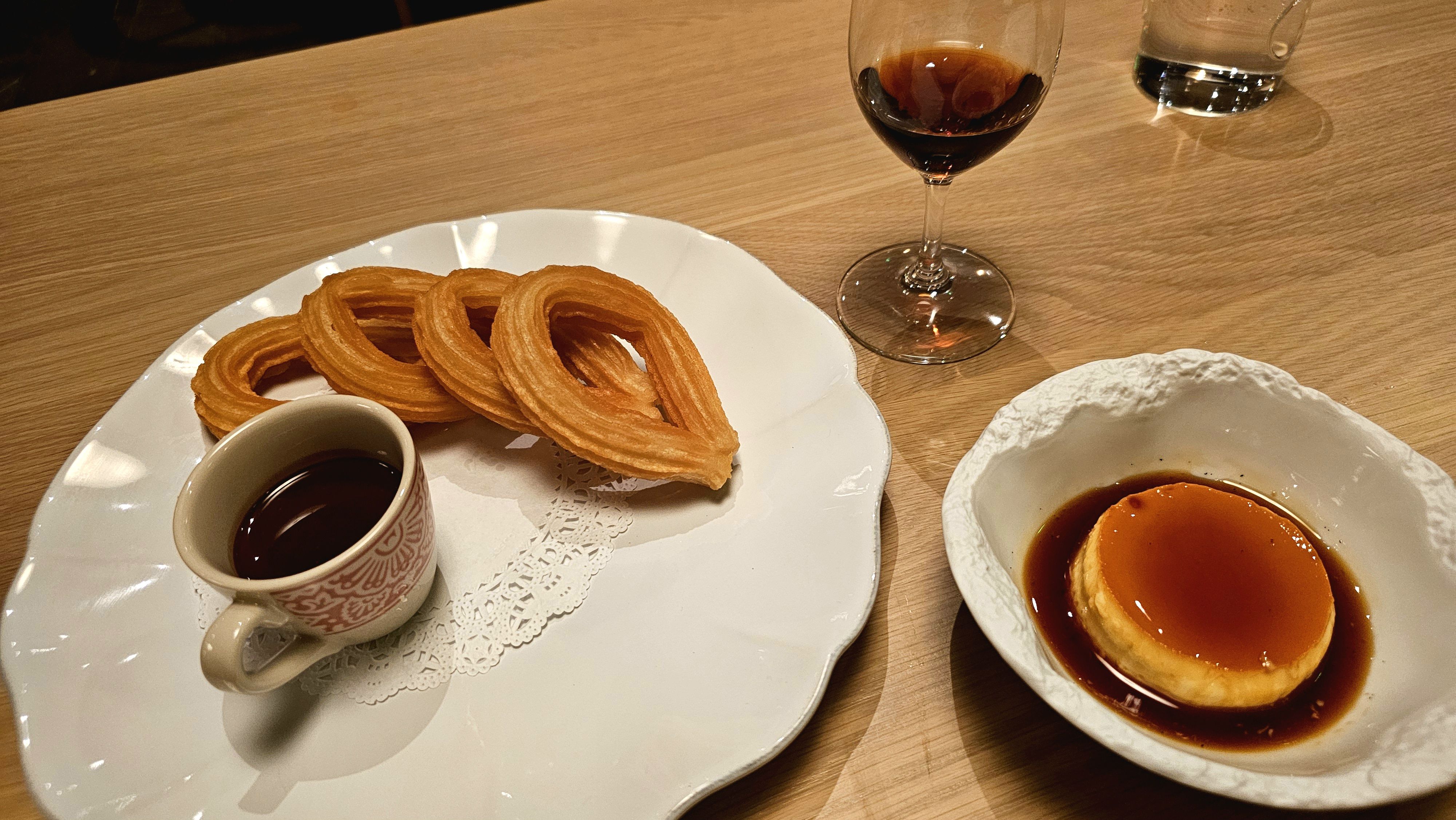 On a wooden table, a white plate holds circular churros with a small cup of dark coffee; a white bowl contains caramel flan in syrup; a glass of red wine sits nearby.