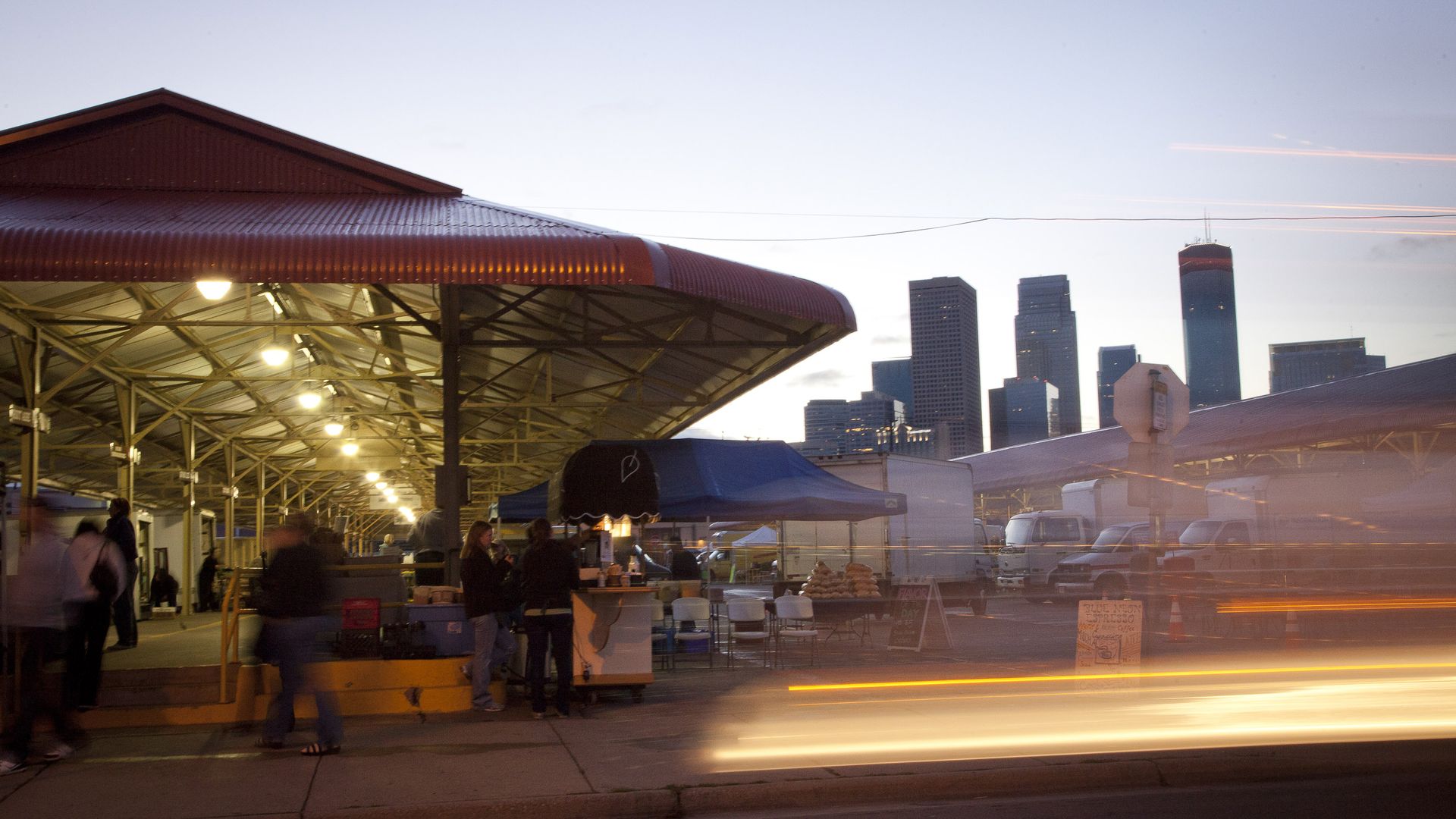 Market scene at dusk beneath a red curved roof with metal framework. Warm lights illuminate stalls and produce; blurred shoppers mingle. In the background, a city skyline rises with light trails along the streets.