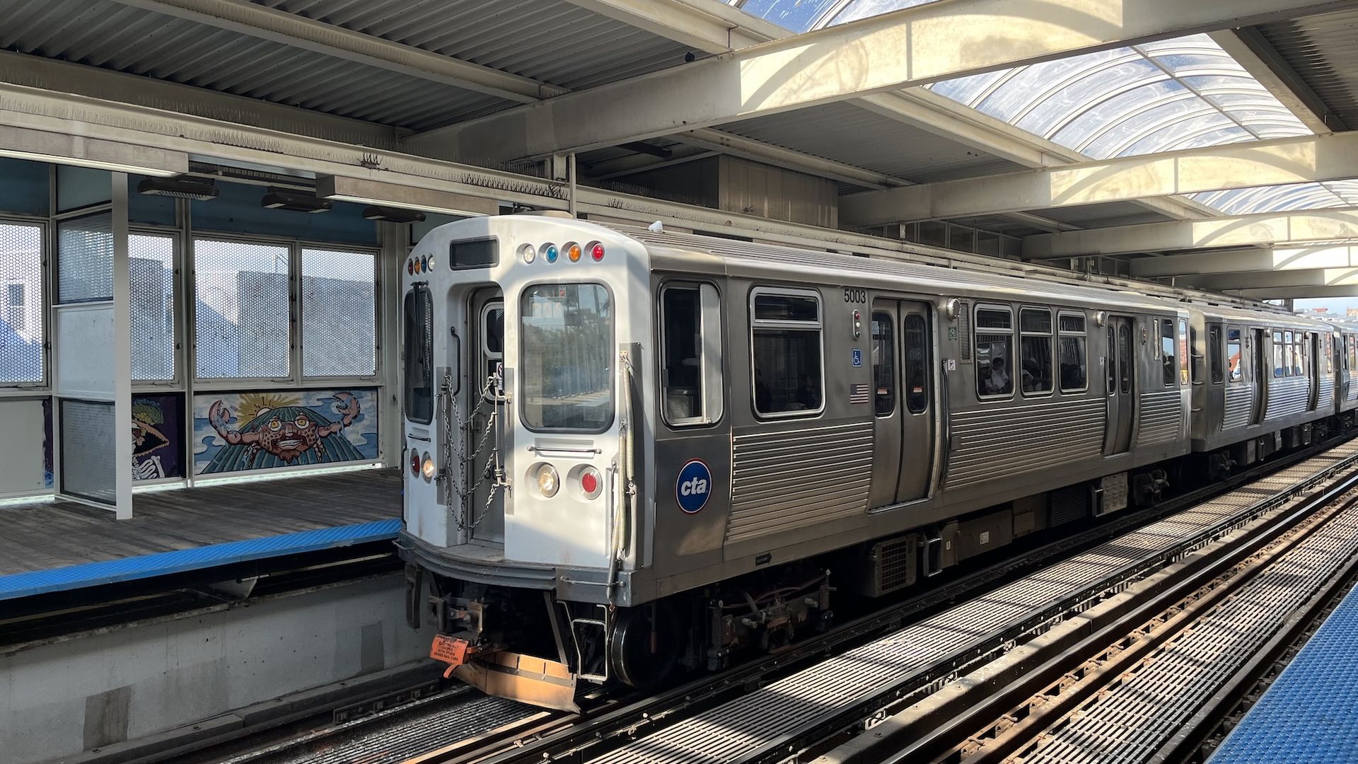 Silver CTA subway train at an elevated station. Doors closed, car 5003, blue platform edge, graffiti mural on the wall, and a curved glass canopy overhead.