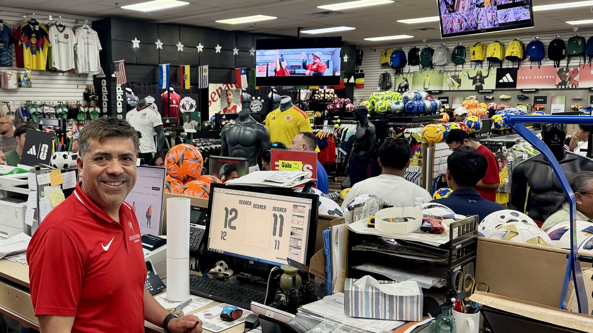 Smiling man in a red polo stands at a busy sports store counter, with jerseys on the wall, backpacks and balls on display, and screens showing sports footage behind the cluttered checkout area.