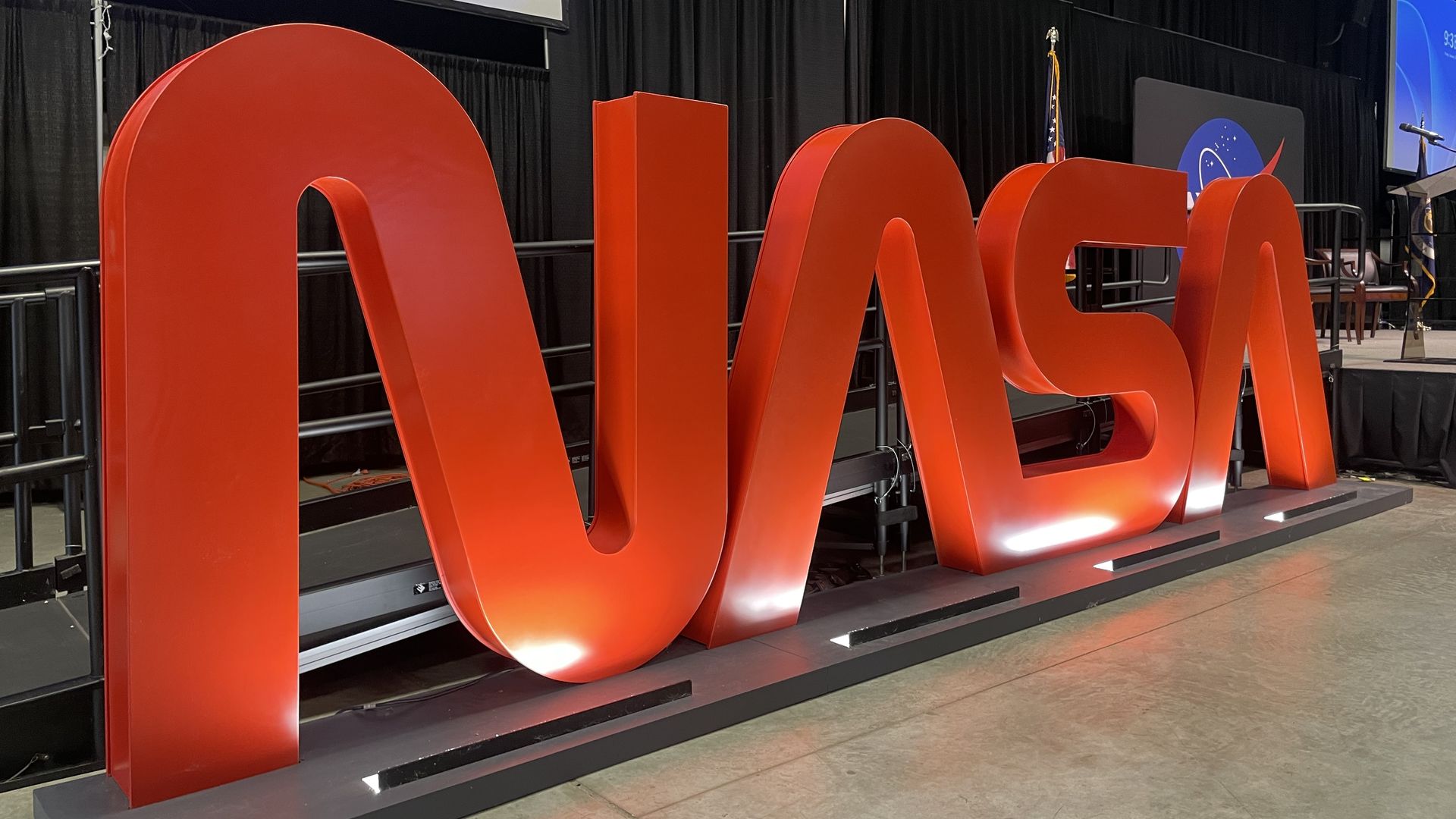 Giant glossy red letters spelling NASA stand on a stage with black curtains, an American flag, and a NASA backdrop; a podium and chairs are visible to the side.