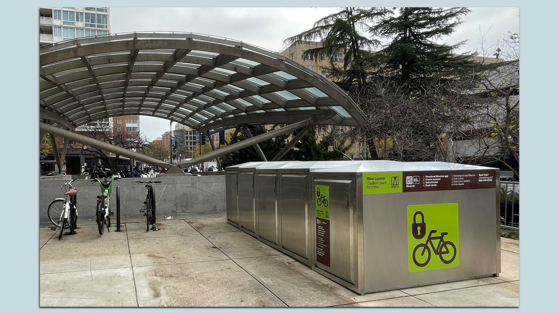 Outdoor bike locker station with silver metal lockers and green bike lock symbols, three bikes parked nearby under a modern curved glass canopy, urban buildings and trees in background.