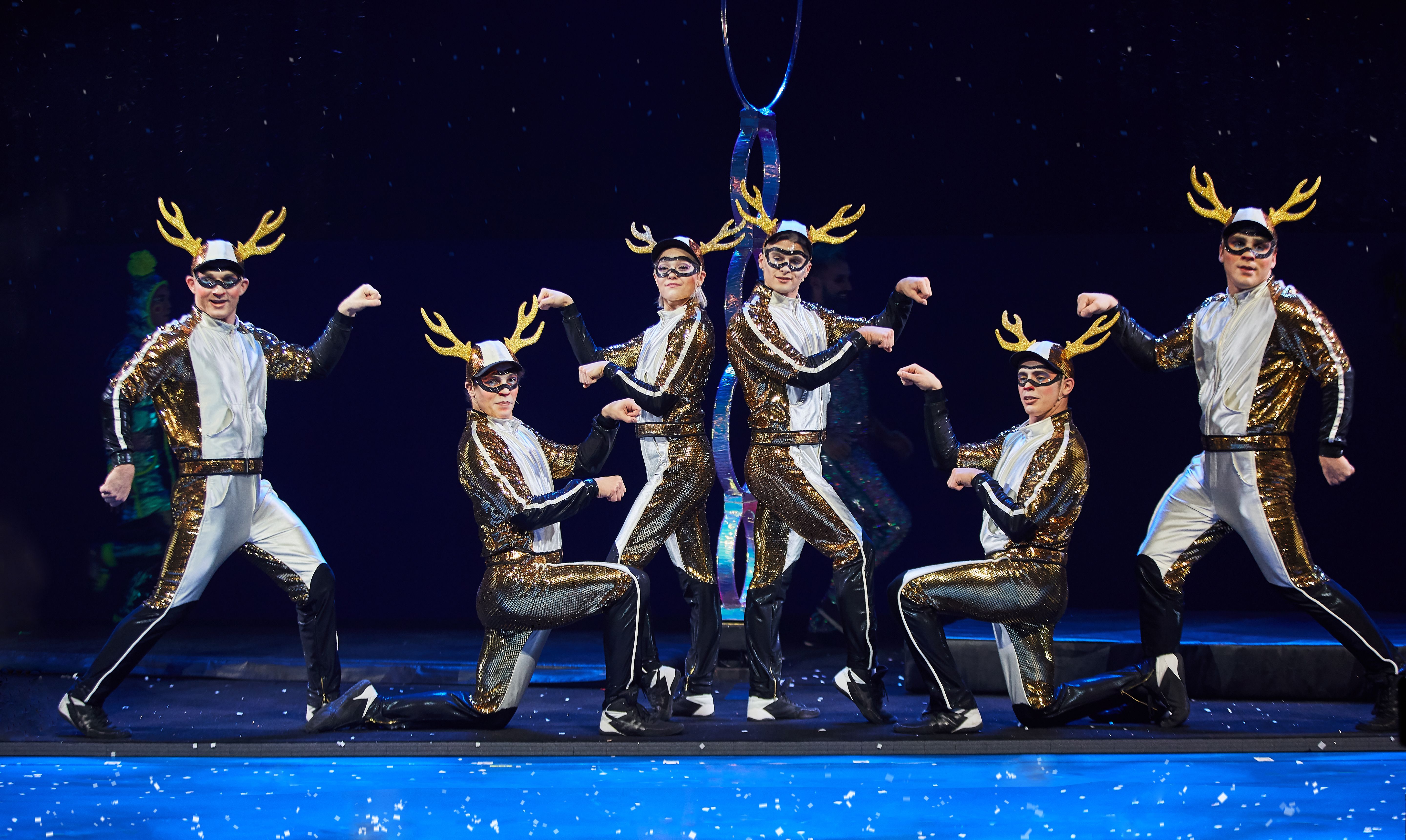 Six performers in gold and black sequined costumes with white fronts, black masks, and golden antlers pose on stage, striking strong arm flexing poses under blue stage lighting.