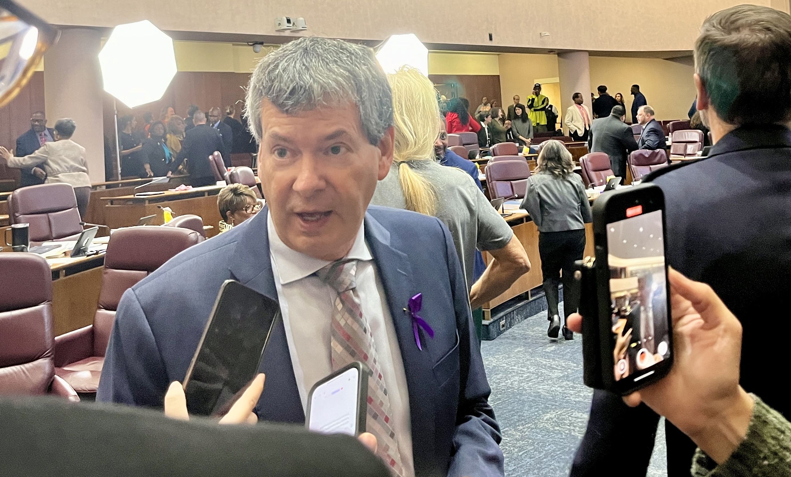 Man in suit with purple ribbon speaks to reporters holding phones in a busy conference or council room with multiple people and bright lights.