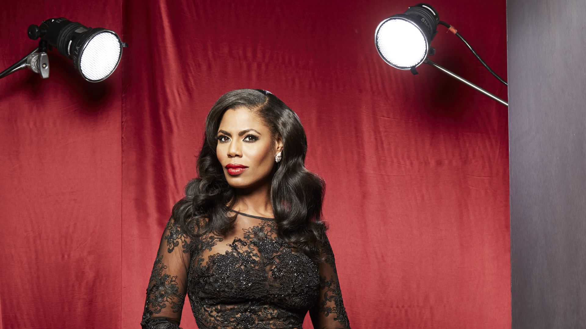 Omarosa standing in front of a red curtain with spotlights on either side, wearing a sexy black dress
